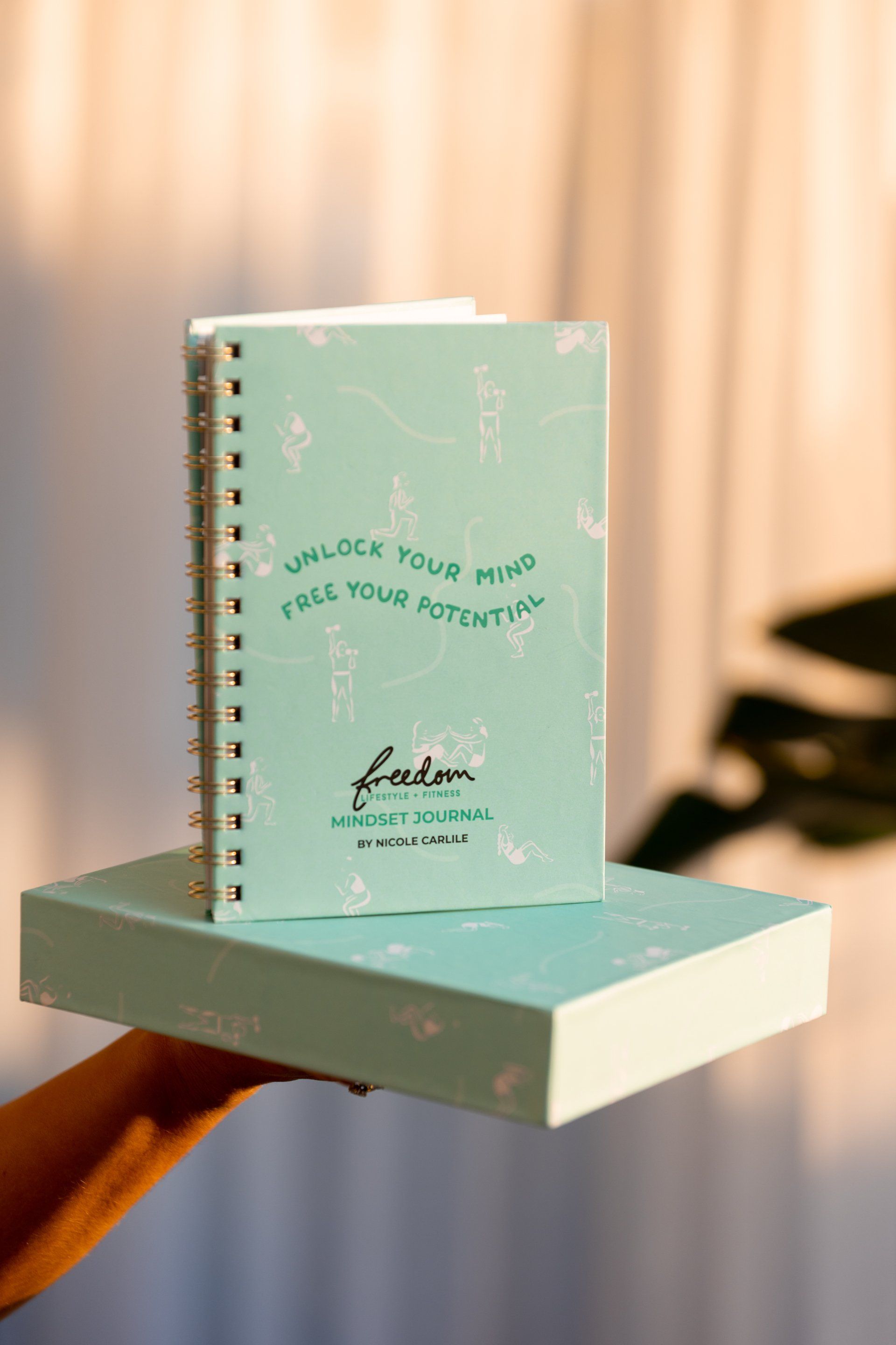 Mint spiral notebook with gold text on a matching box against a blurred curtain backdrop