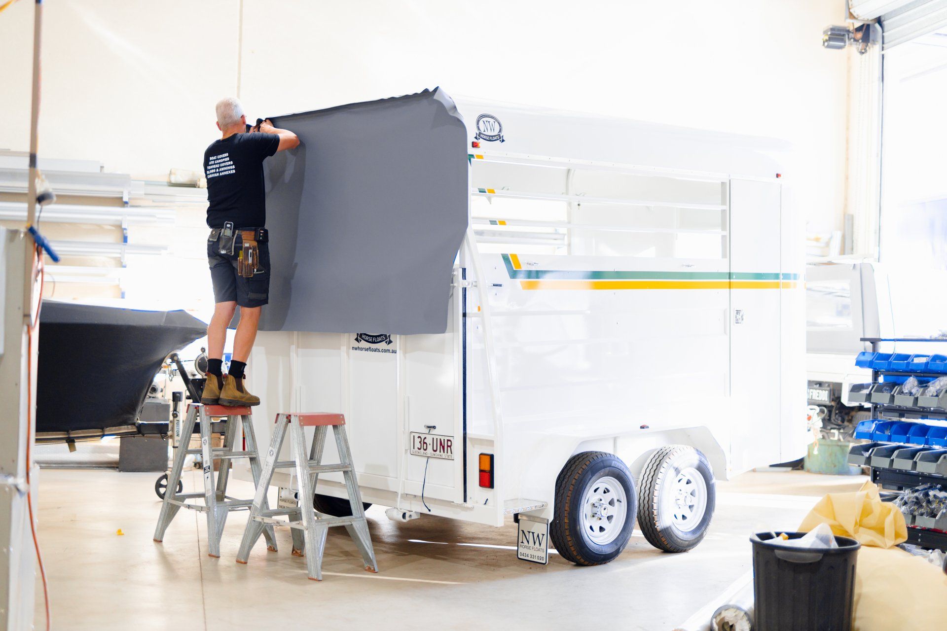 Worker on a ladder installing a large gray panel on a white trailer in a bright workshop