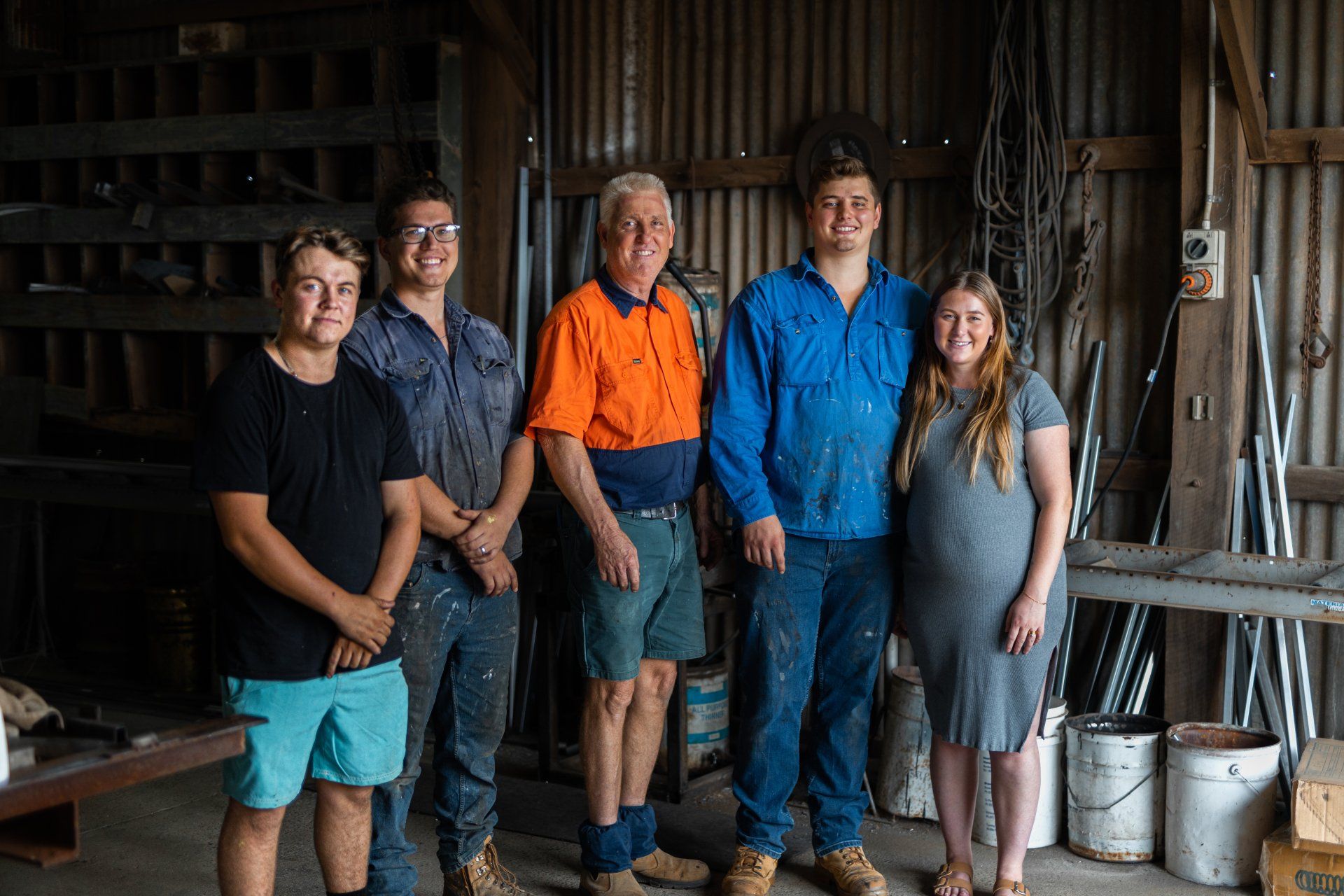 Five people stand smiling in a rustic workshop, with tools, shelves, and buckets in the background.