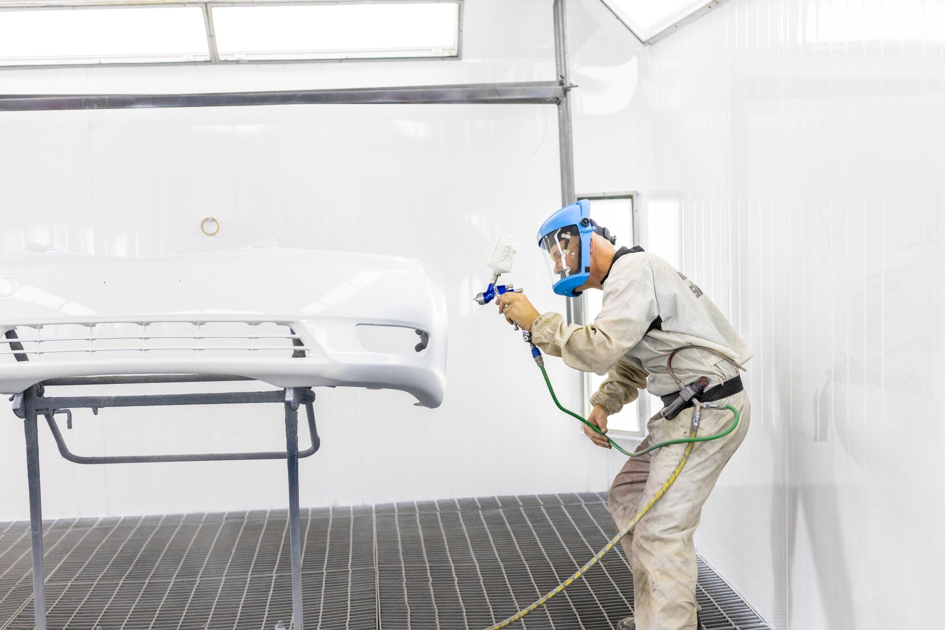 Worker in protective gear spray-painting a car body in a white auto paint booth