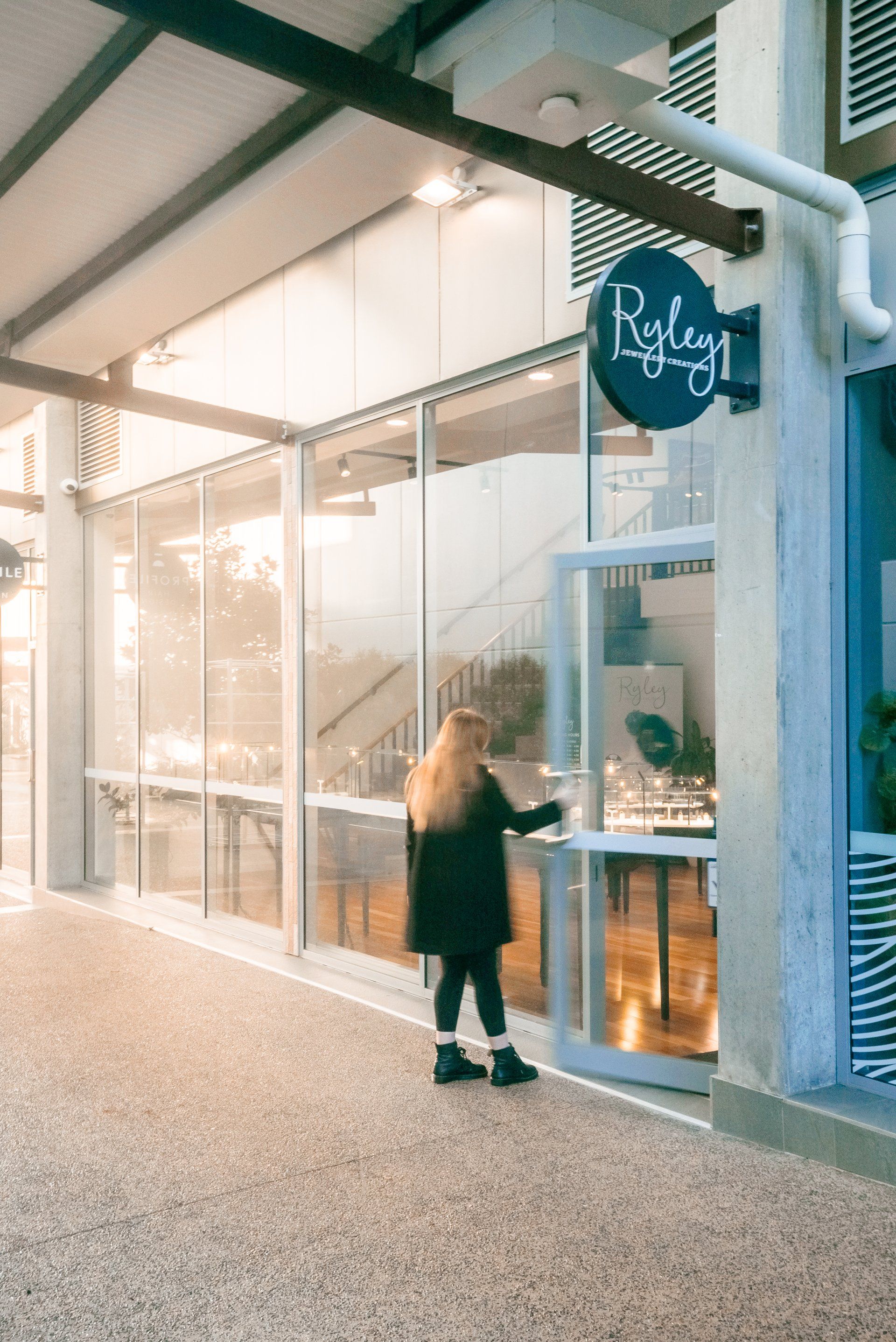 Person standing at a glass storefront counter, reaching toward the window under a round hanging sign.