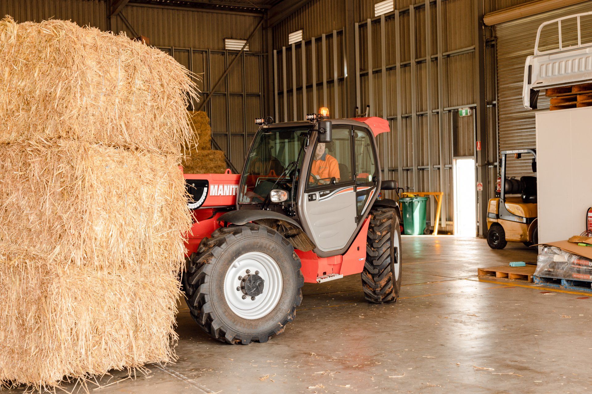 Red tractor inside a barn beside a large hay bale, with a forklift in the background.