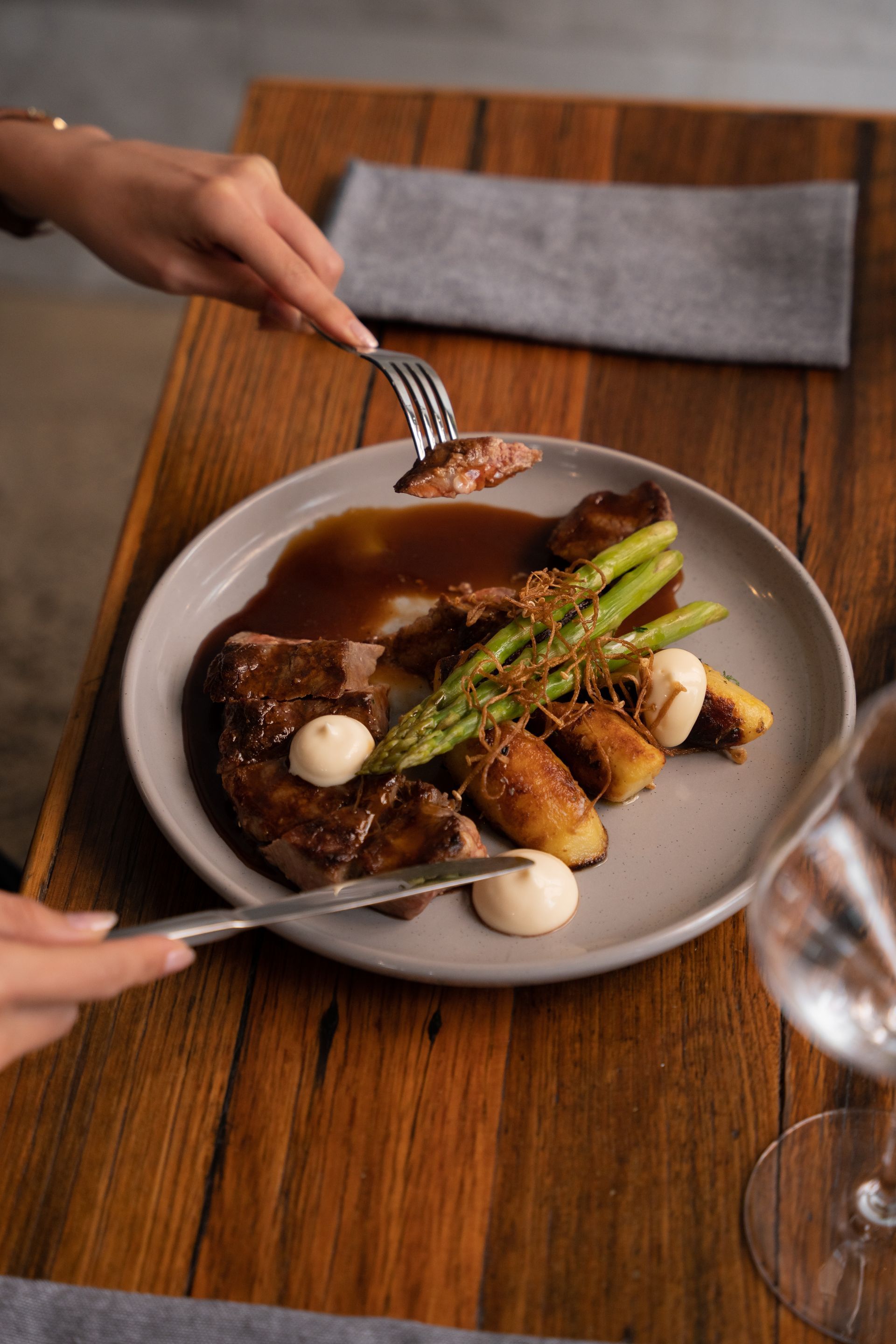 Hands serving a plated steak with asparagus and sauce on a dining table