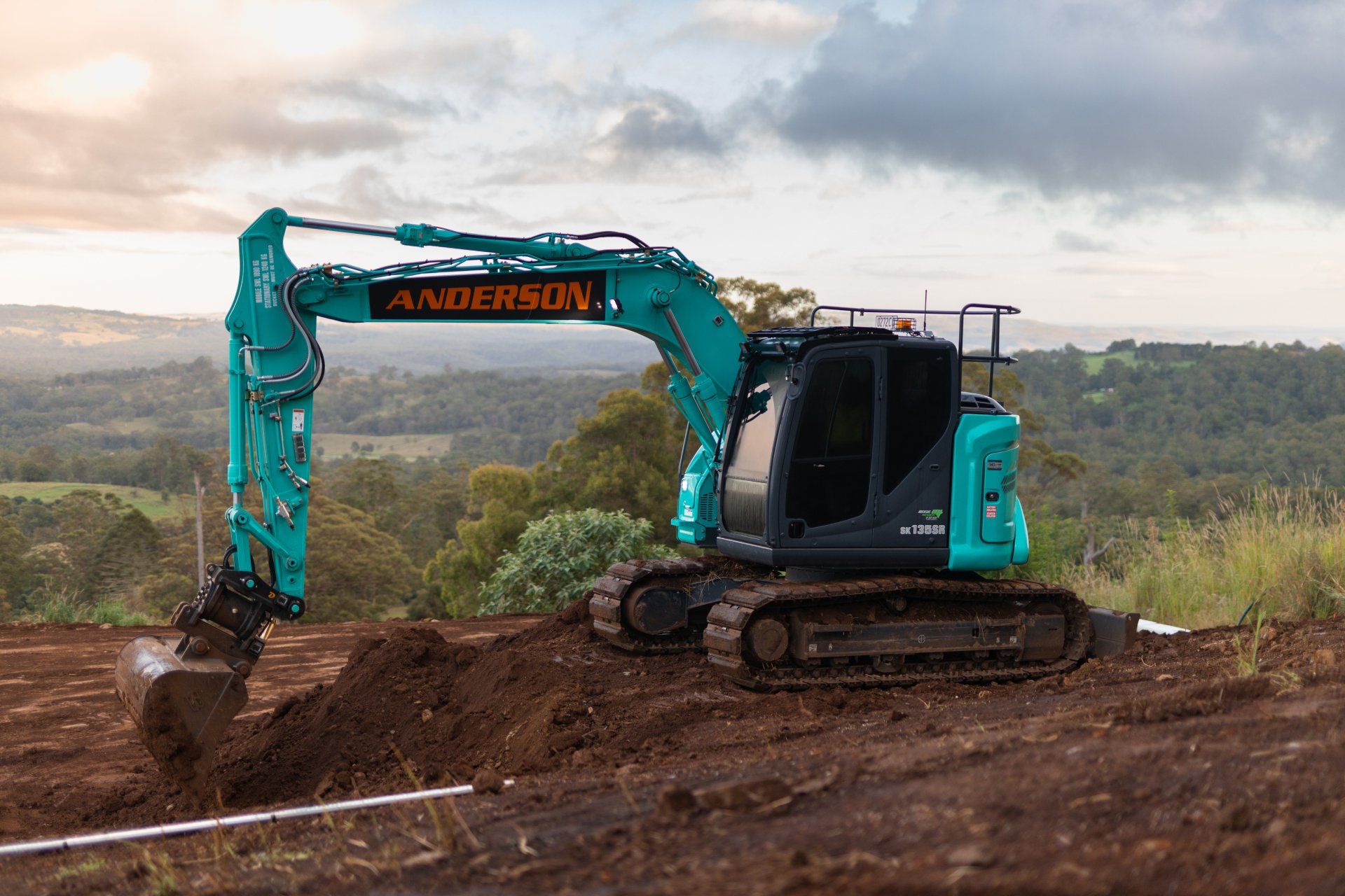 Turquoise excavator digging dirt on a hillside with a forested landscape in the background