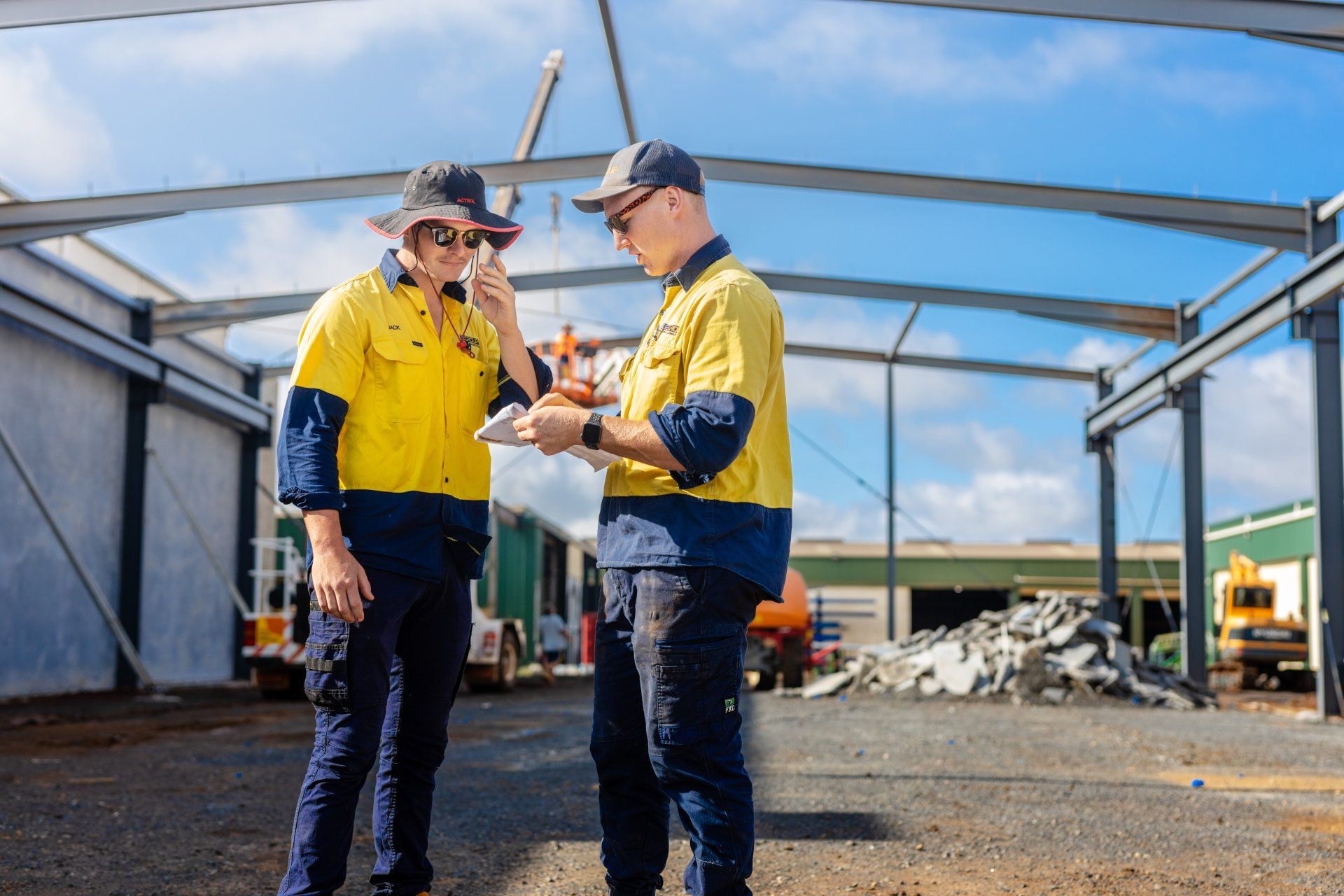 Two workers in yellow jackets discussing plans at a construction site with a steel frame and rubble.