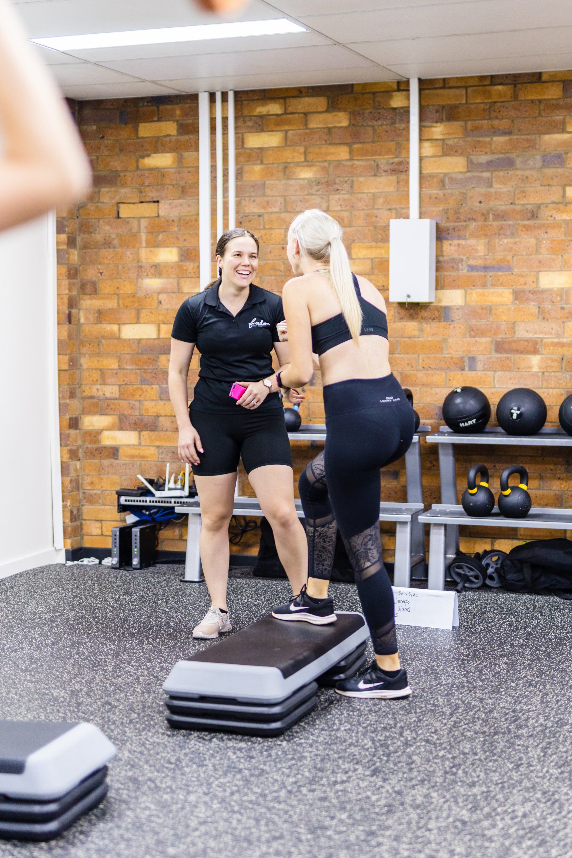 Two women exercising in a gym, one stepping on a platform while talking with the other