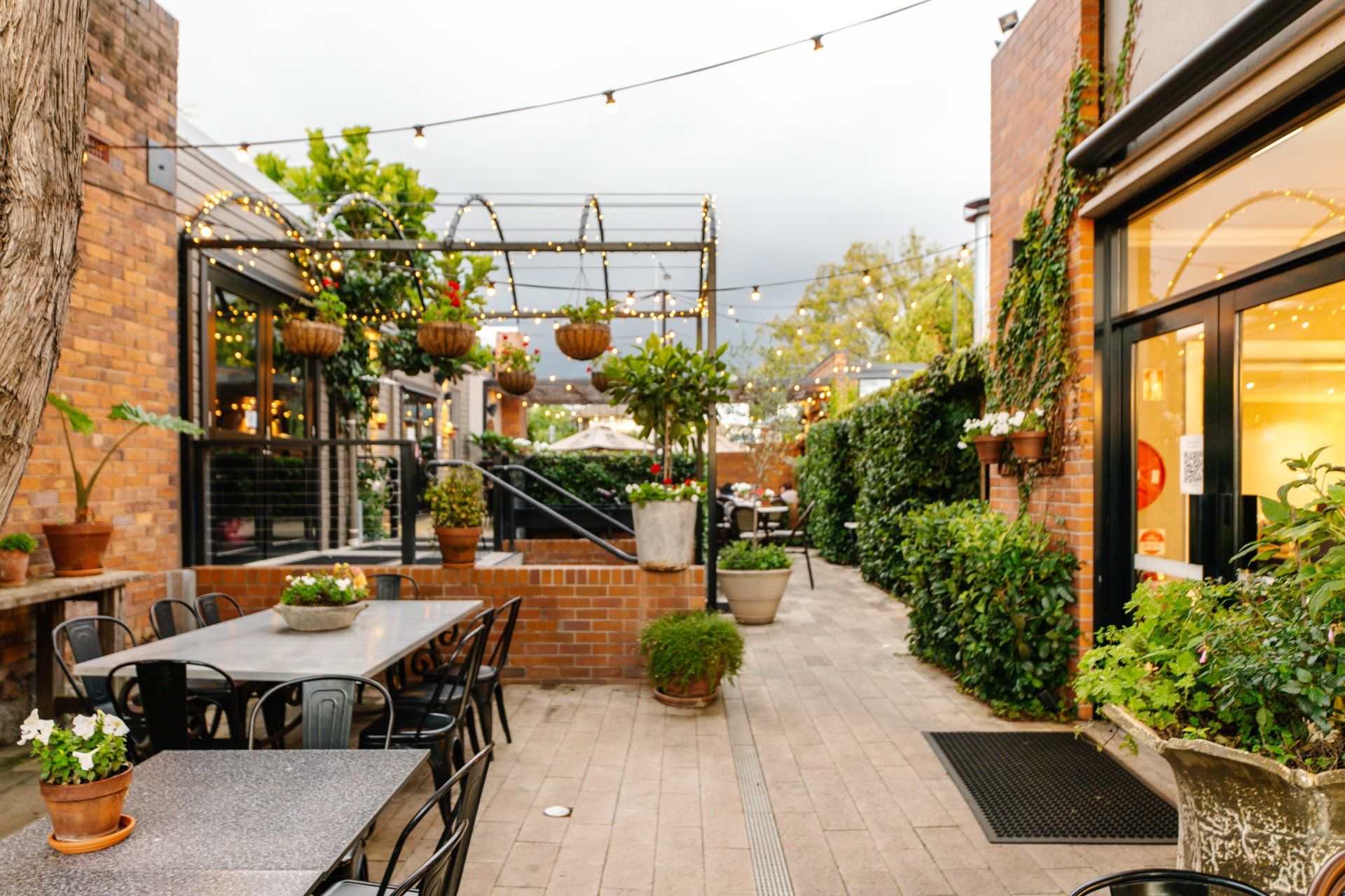 Outdoor café patio with tables, chairs, potted plants, and string lights along a brick walkway