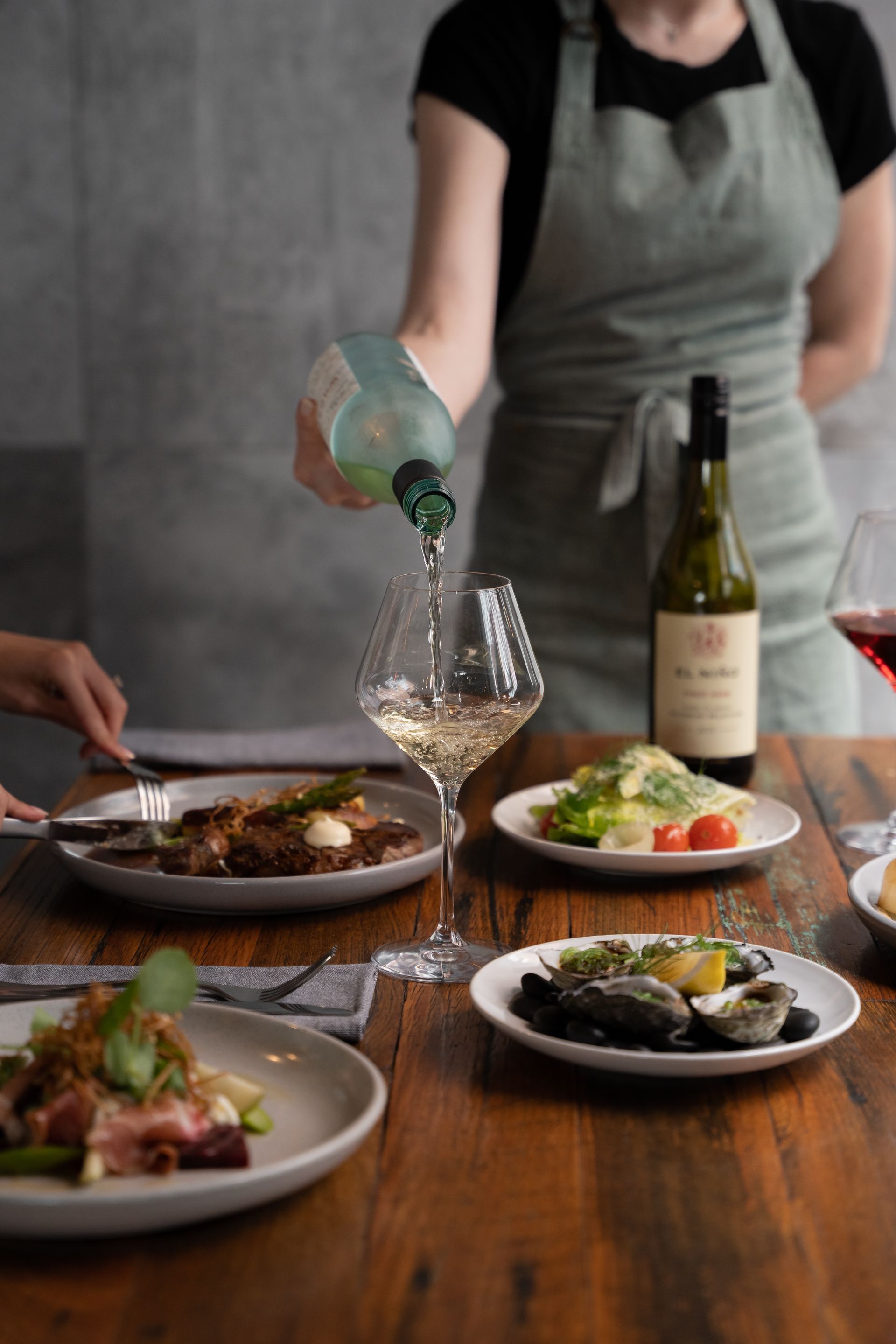Server pouring white wine at a restaurant table with plated dishes and a wine bottle