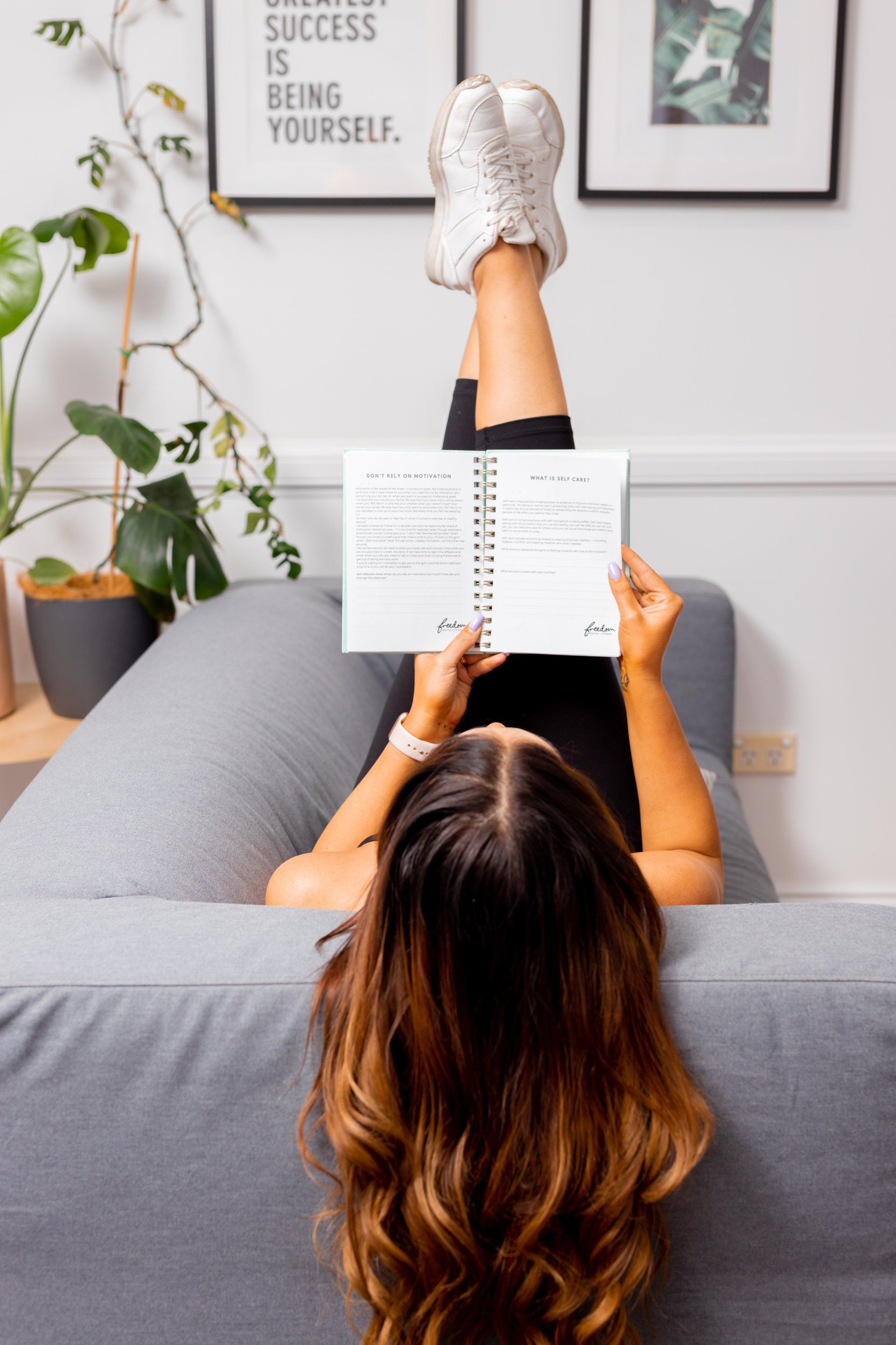 Person lying on a gray couch reading a notebook in a bright living room with plants and wall art