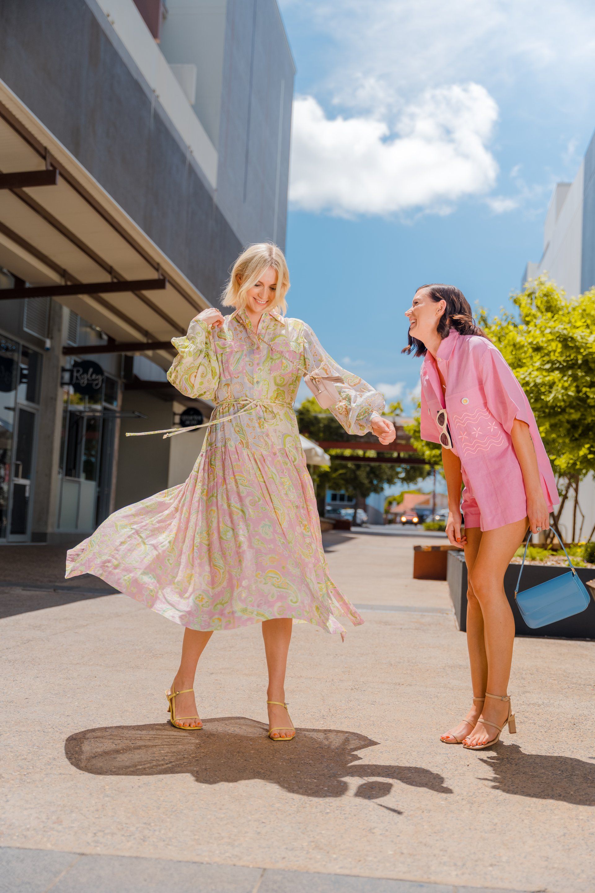 Two women in colorful dresses chatting outdoors on a sunny walkway near modern buildings.