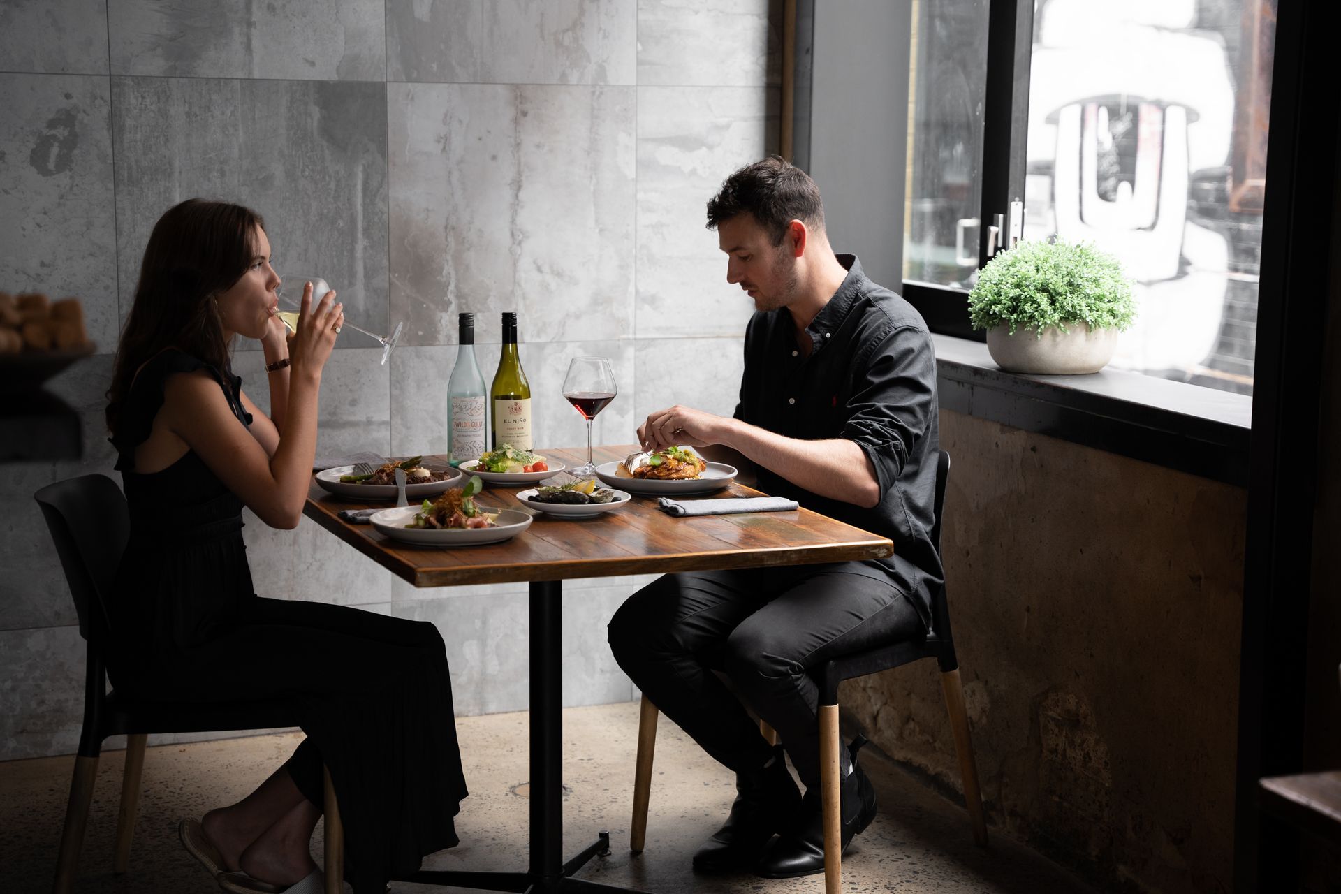 Two people eating and talking at a small table in a cozy restaurant by a window