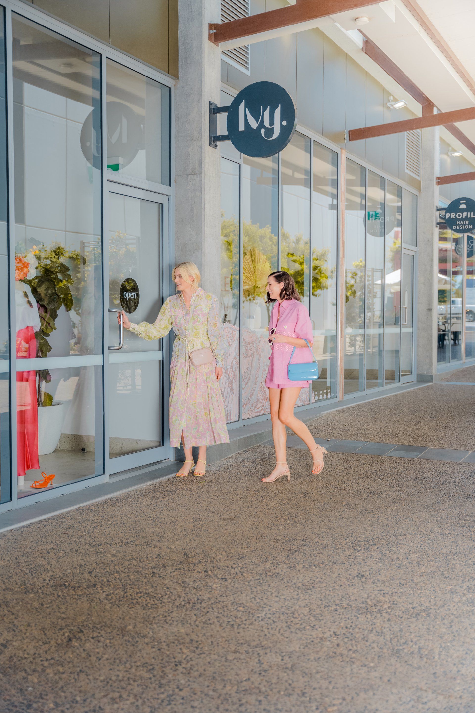 Two women in pastel outfits walk past glass storefronts under a covered walkway.