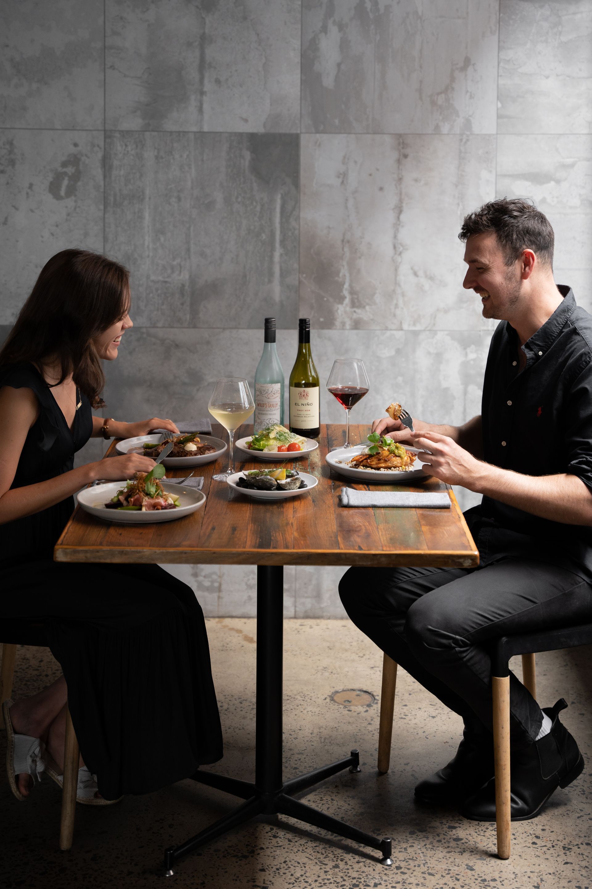Two diners face each other at a small restaurant table with plated meals and wine against a gray wall.