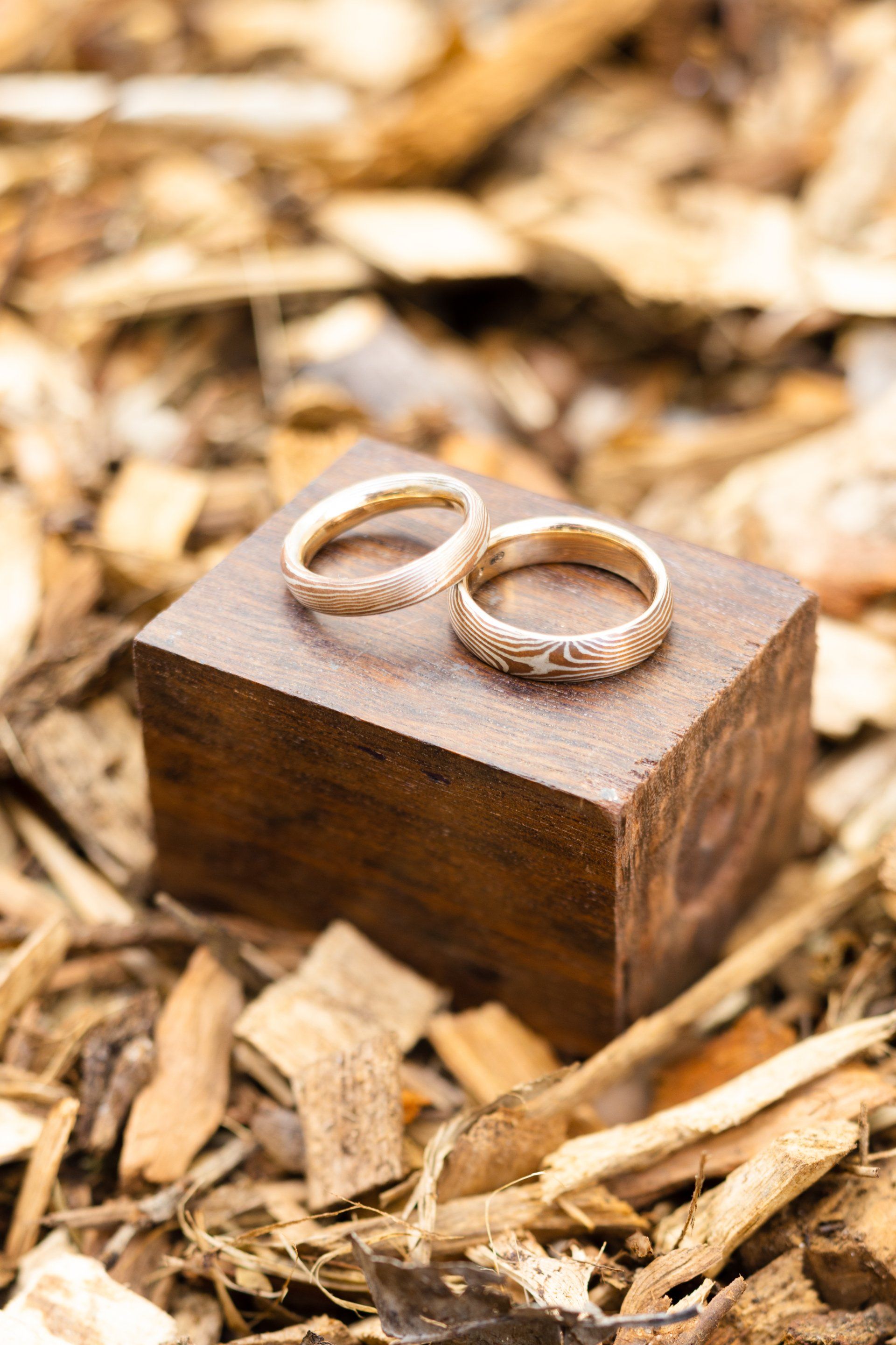 Two gold rings on a small wooden block amid dry leaves and wood chips