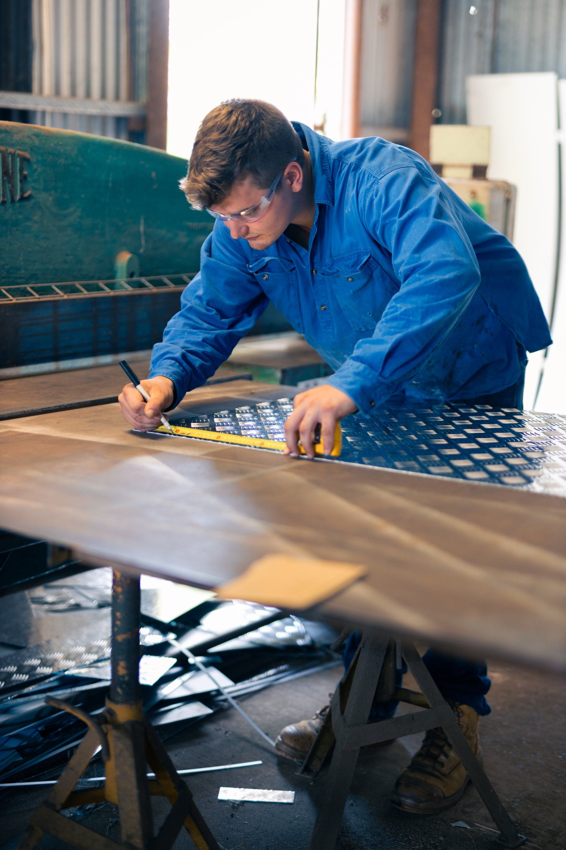 Worker in blue coveralls measuring and marking a large metal sheet in a workshop.