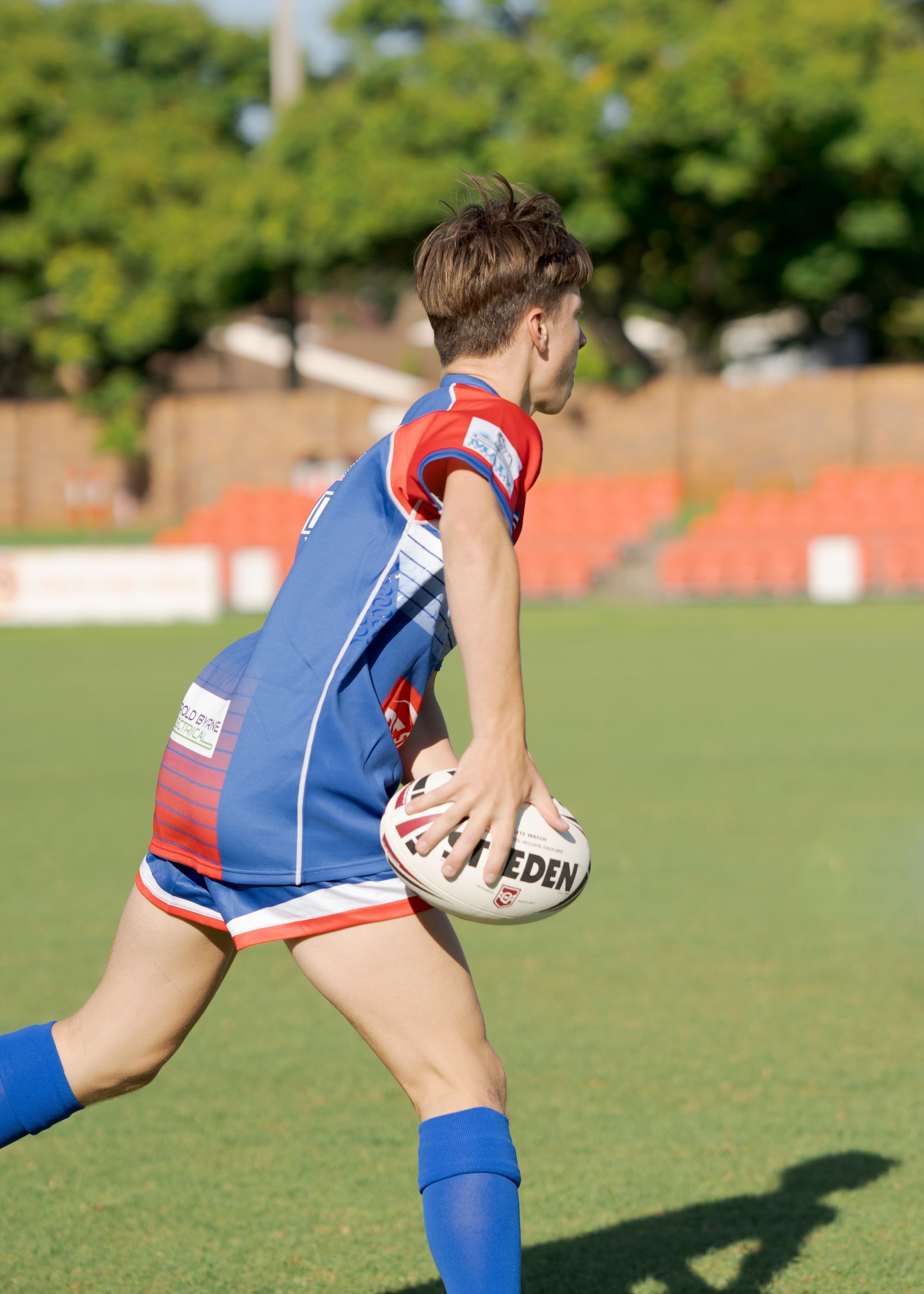 Rugby player in blue and red uniform running with the ball on a grassy field