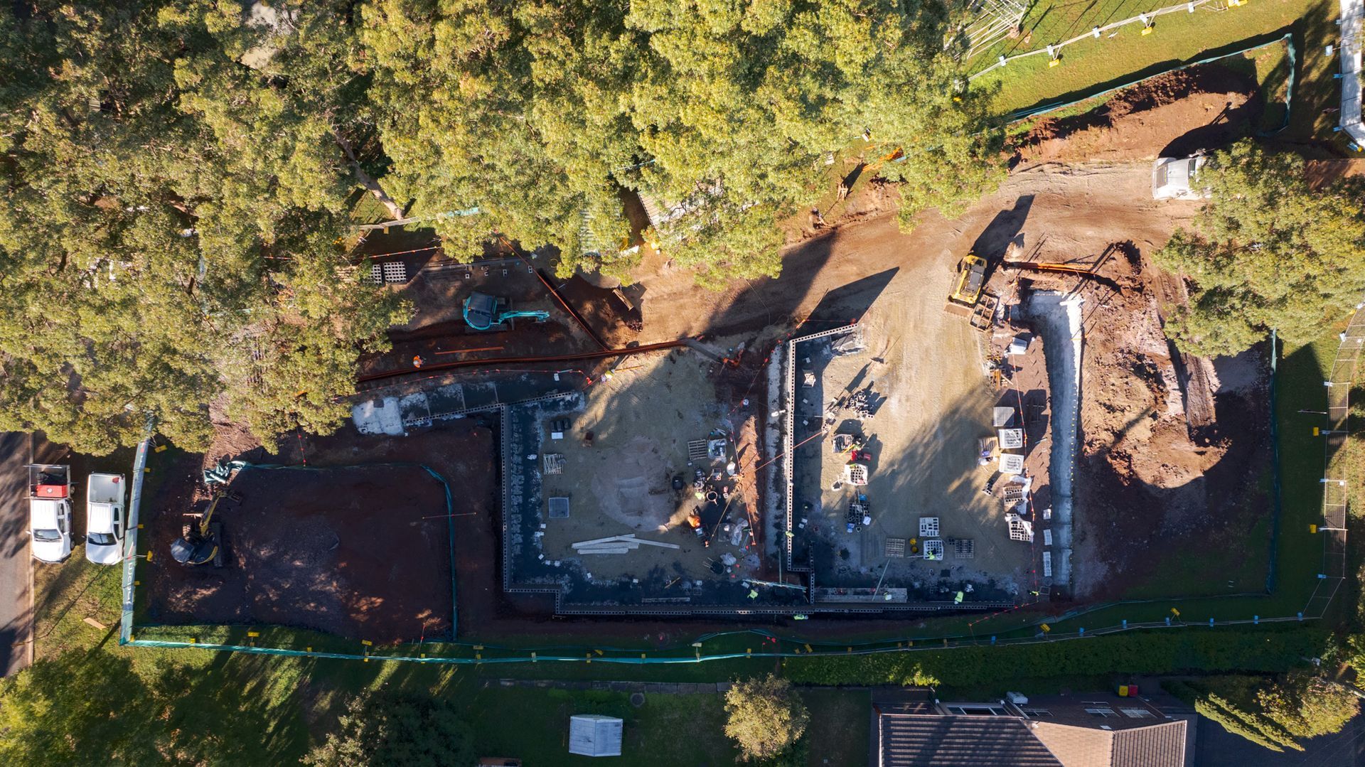 Aerial view of a construction site with excavation, dirt piles, and building materials beside trees and a road
