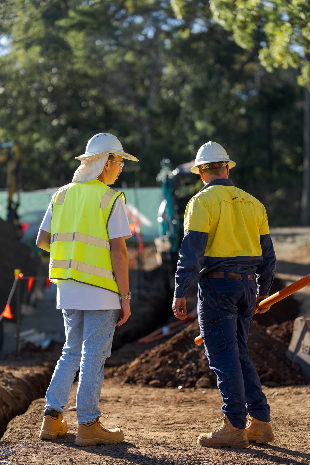 Two construction workers in hard hats and safety vests talking at a roadwork site.