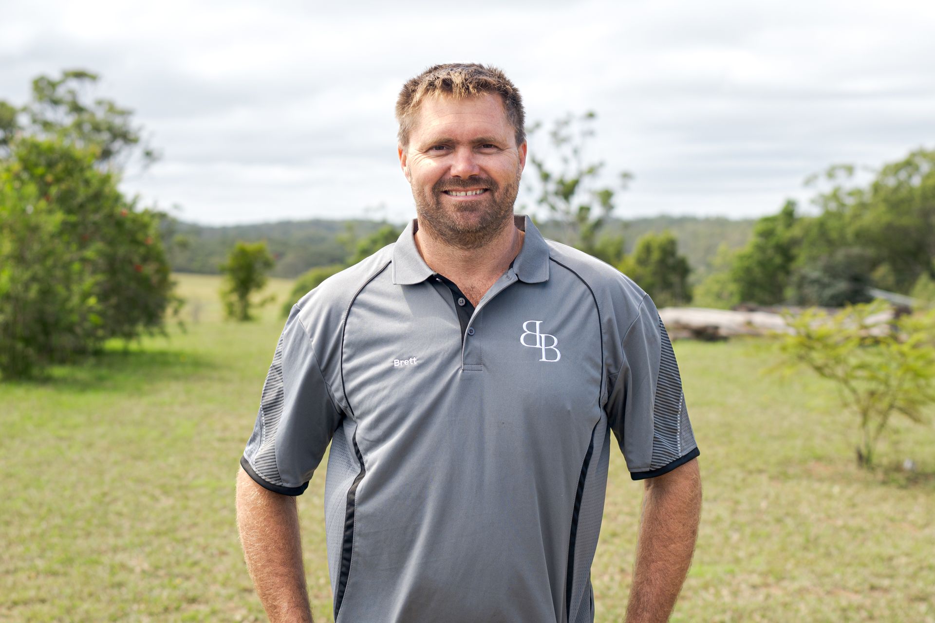 Man standing outdoors on a grassy field, wearing a gray polo shirt and smiling at the camera.