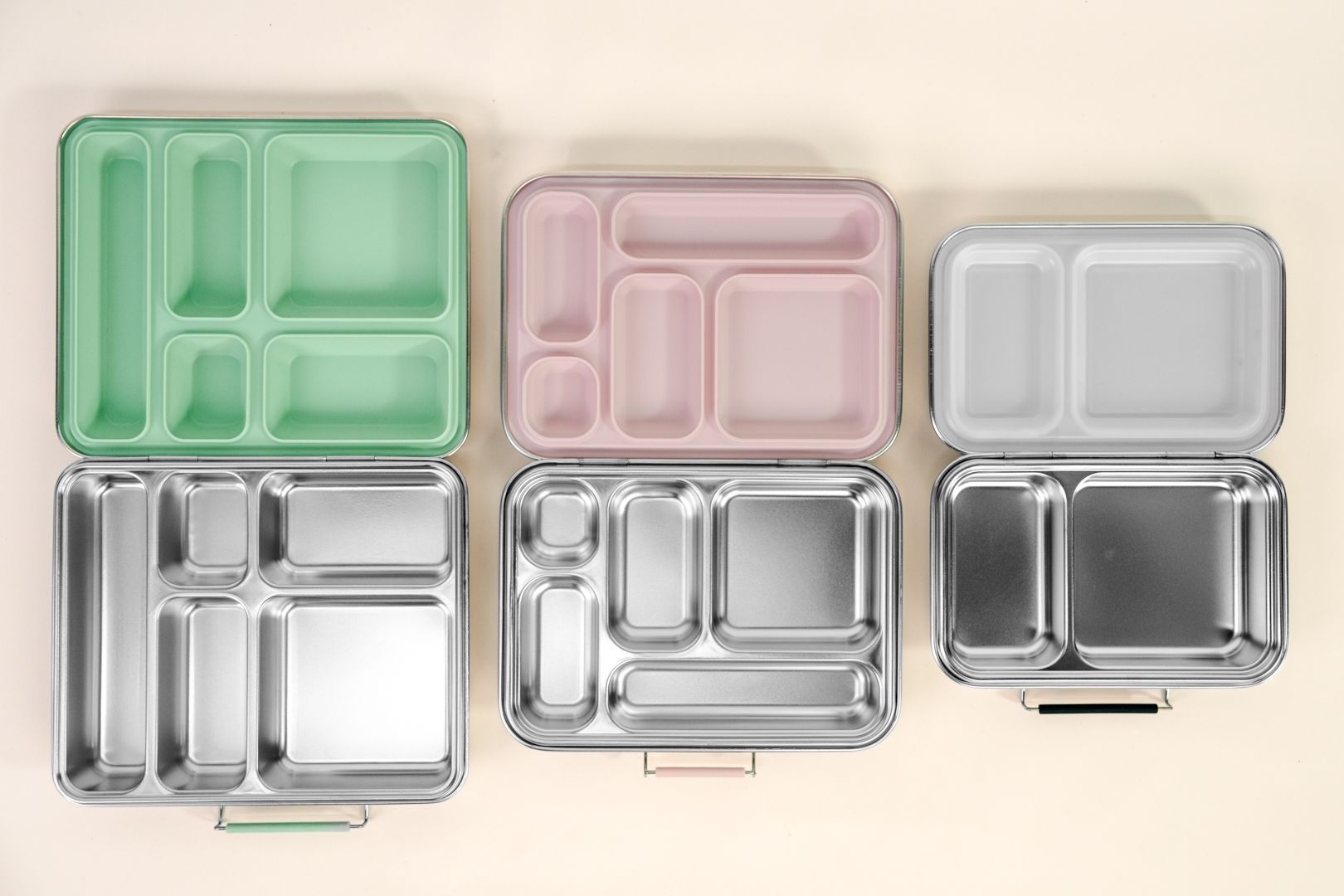 Five compartment lunch boxes in green, pink, gray, and clear plastic on a light background
