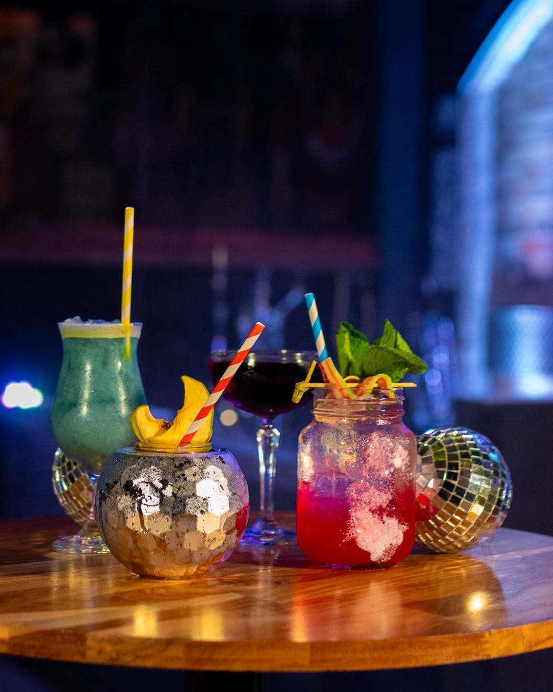 Three colorful cocktails with garnishes on a wooden table in a dimly lit bar setting