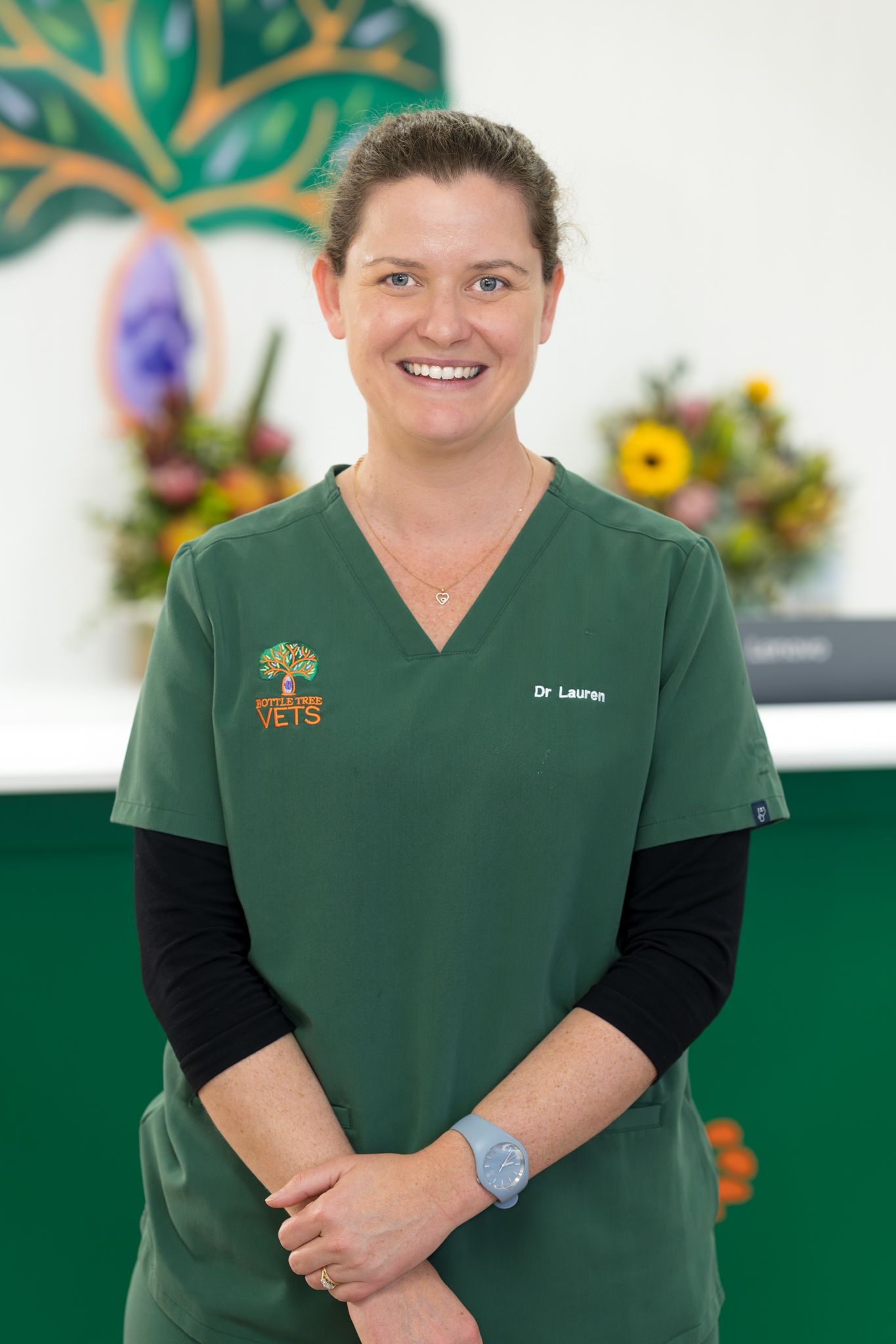 Smiling person in green scrubs standing in a clinic, with a plant mural and flowers in the background