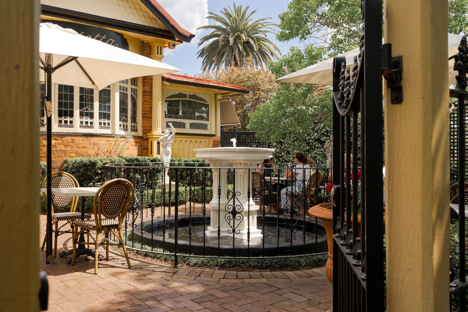 Courtyard with a small fountain, black iron fence, and tan building with palm trees and umbrellas