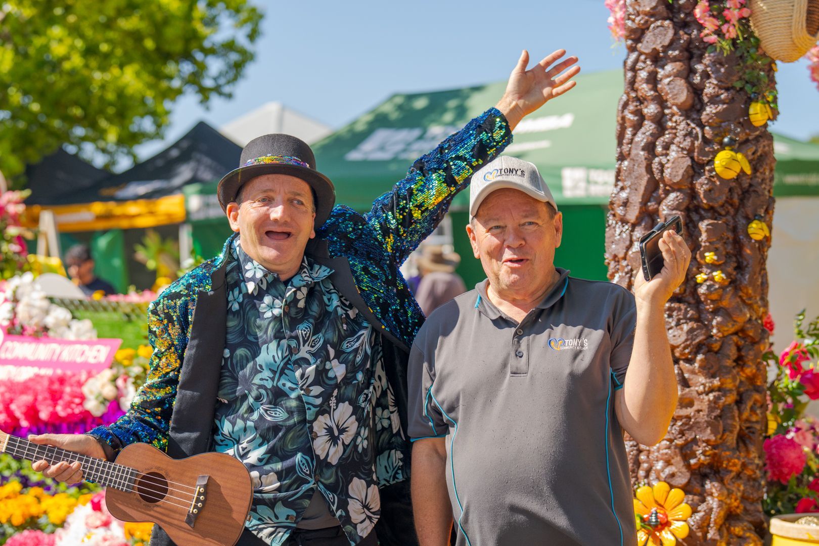 Two smiling people standing by a decorated tree at an outdoor flower market, one waving with an acoustic guitar.