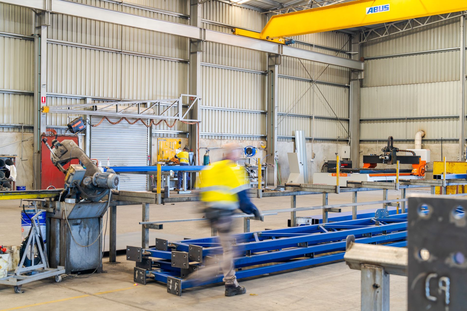 Workers in a metal workshop moving long blue steel beams near machinery and a yellow overhead crane.