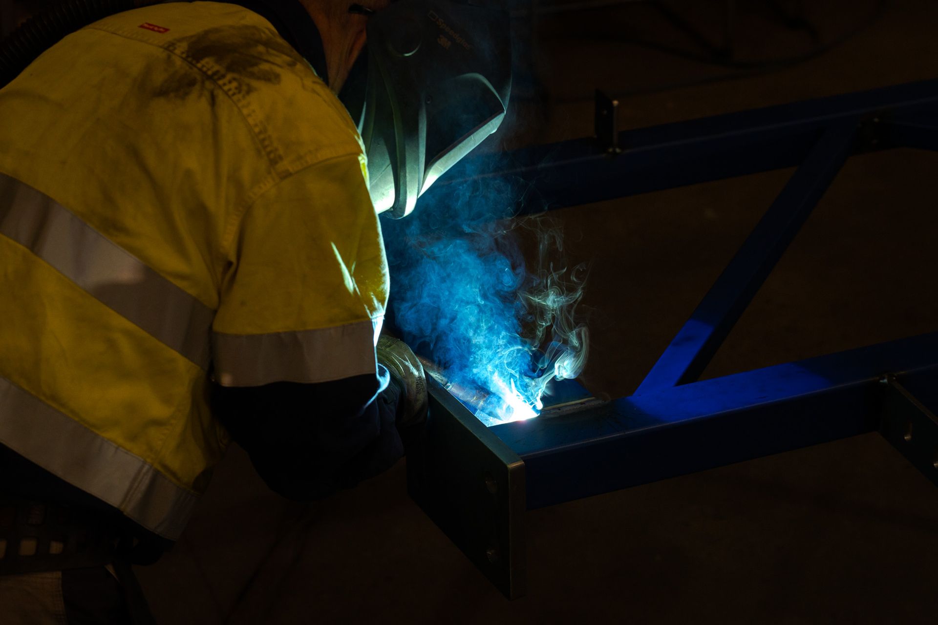 Worker welding metal in a dark workshop, bright blue sparks and smoke visible