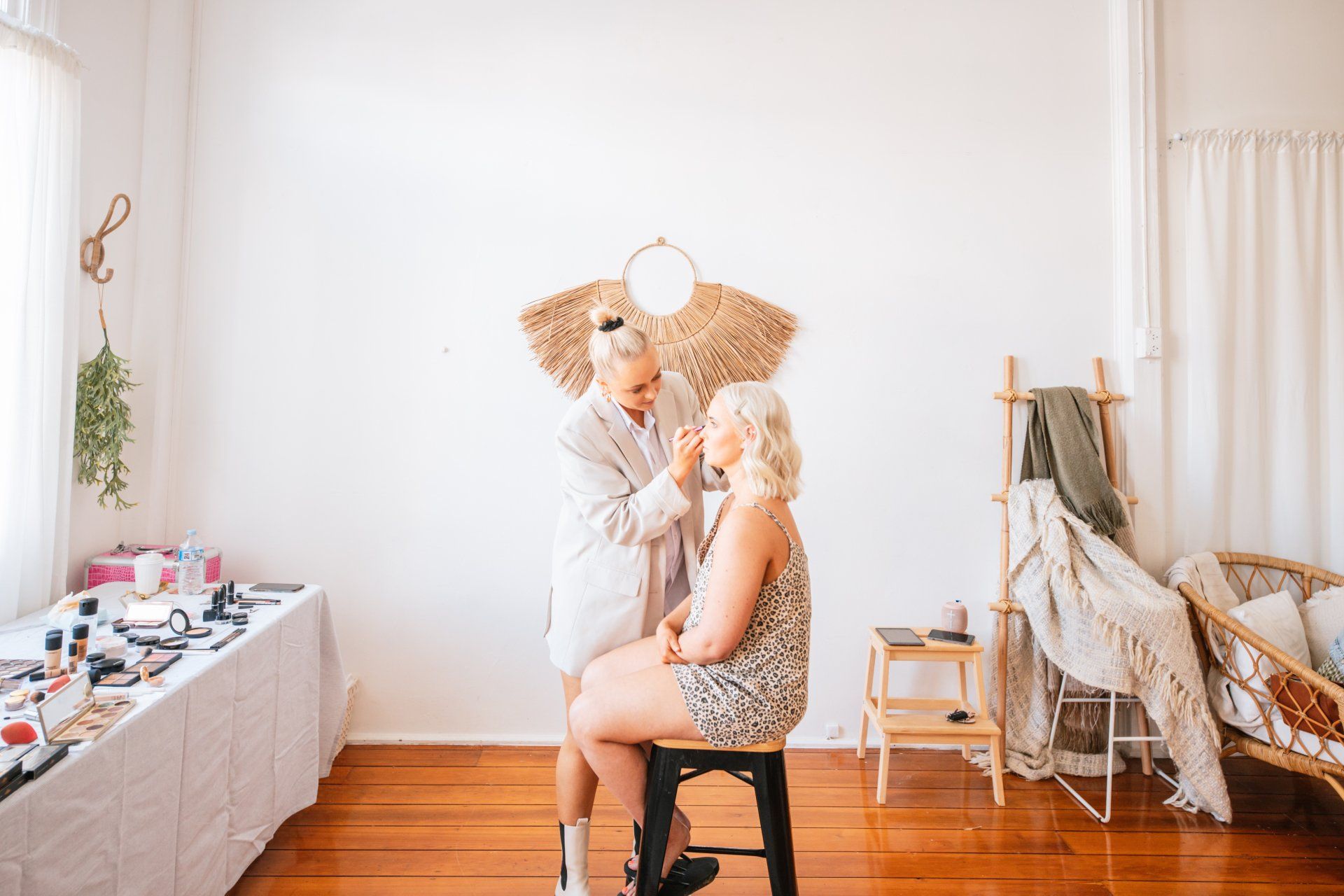 Person styling another seated person in a bright room with boho decor and a table of makeup supplies