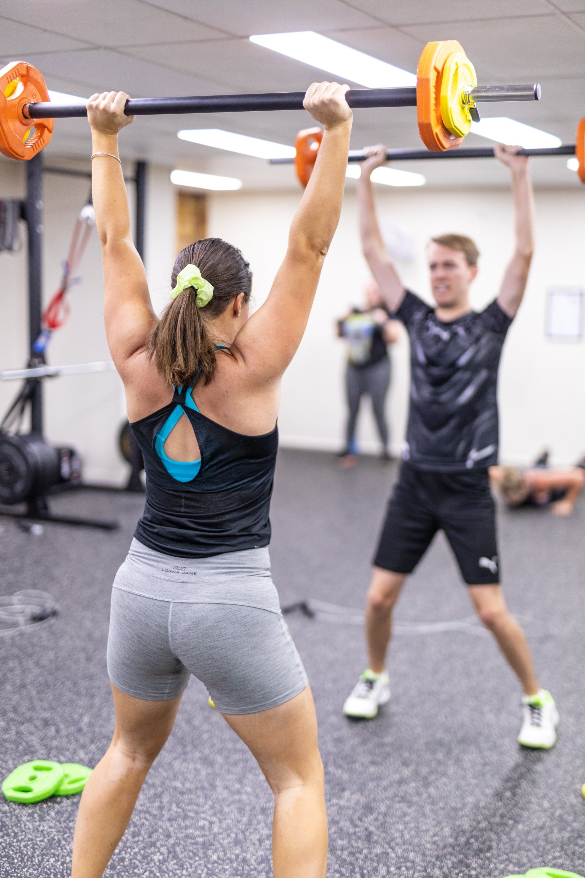 Two people doing overhead barbell lifts in a gym, one facing away in the foreground.