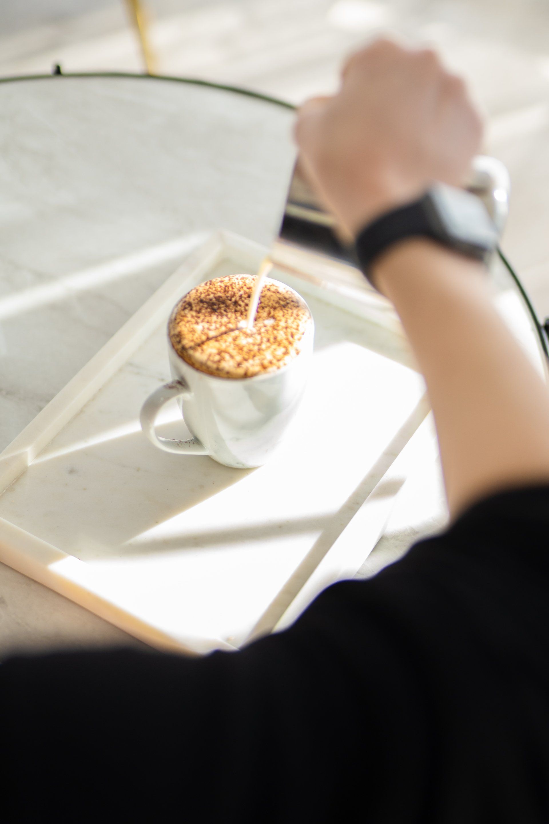 Person stirring coffee on a sunlit white table with a spoon, viewed from above