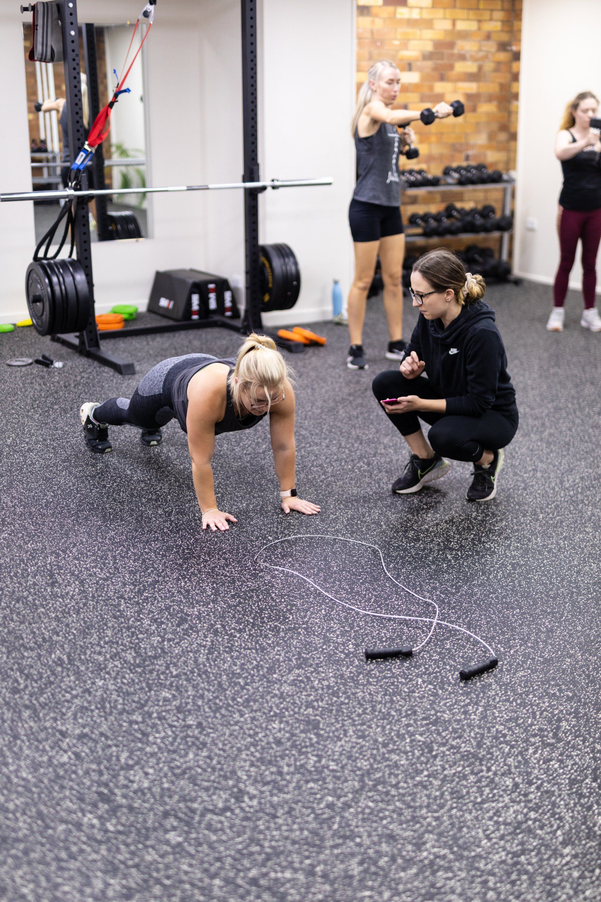 Woman doing push-ups while a coach crouches nearby in a gym with other people in the background
