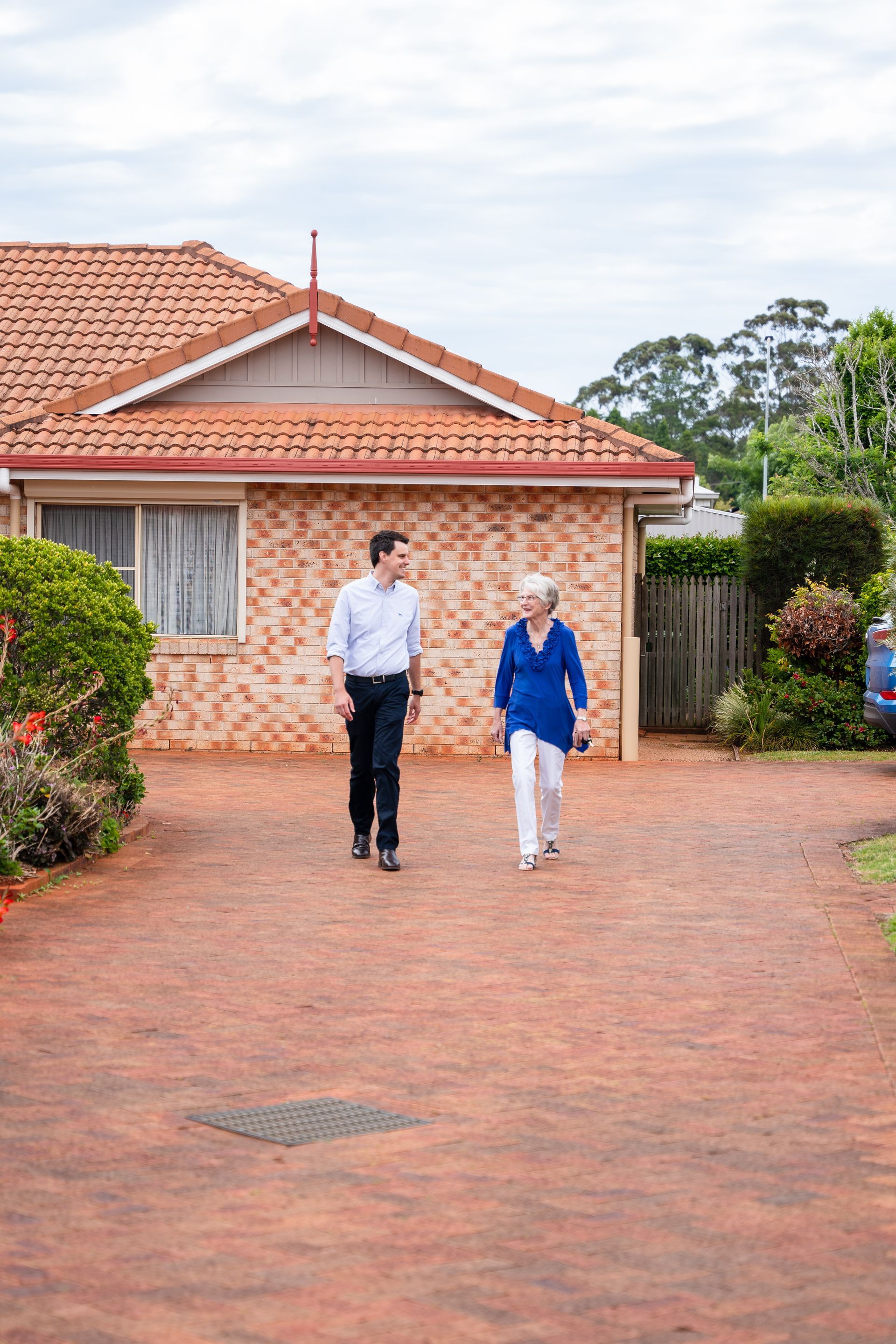 Two people walking on a brick driveway in front of a house with a red-tiled roof