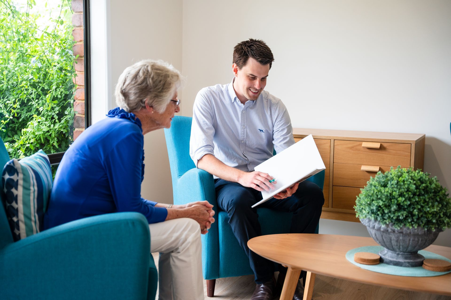 Two people talking in a bright living room, one holding a document beside a coffee table and plant.