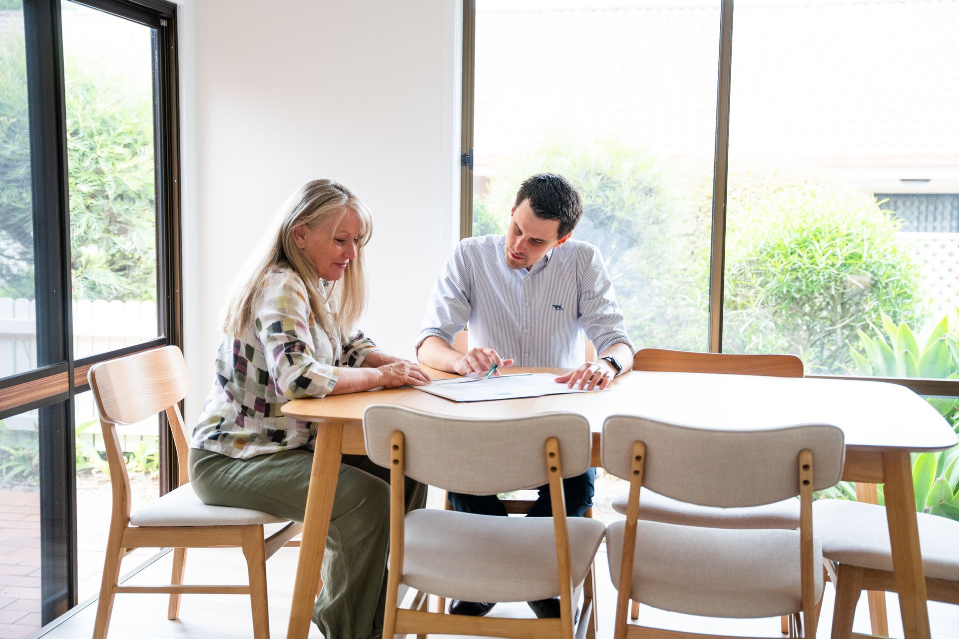 Two people discussing papers at a table in a bright room with large windows.
