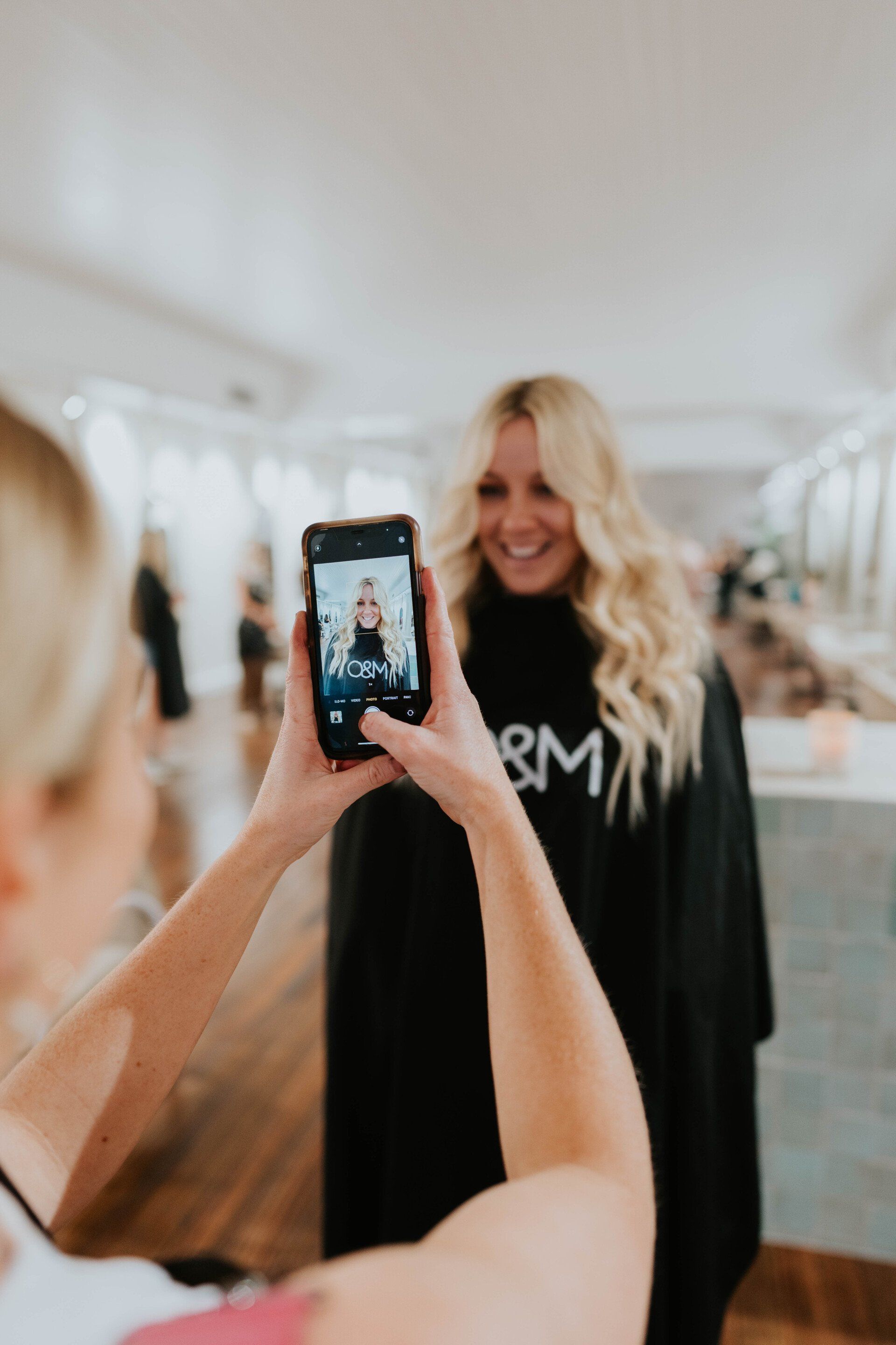 Person taking a photo of a smiling woman with long blonde hair in a bright salon setting