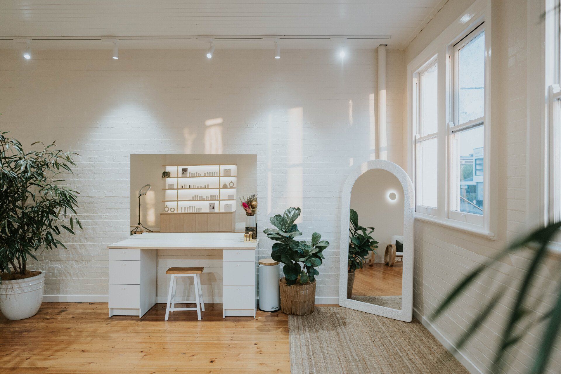 Bright minimalist studio with a white desk, mirror, plants, and large windows in a sunlit room