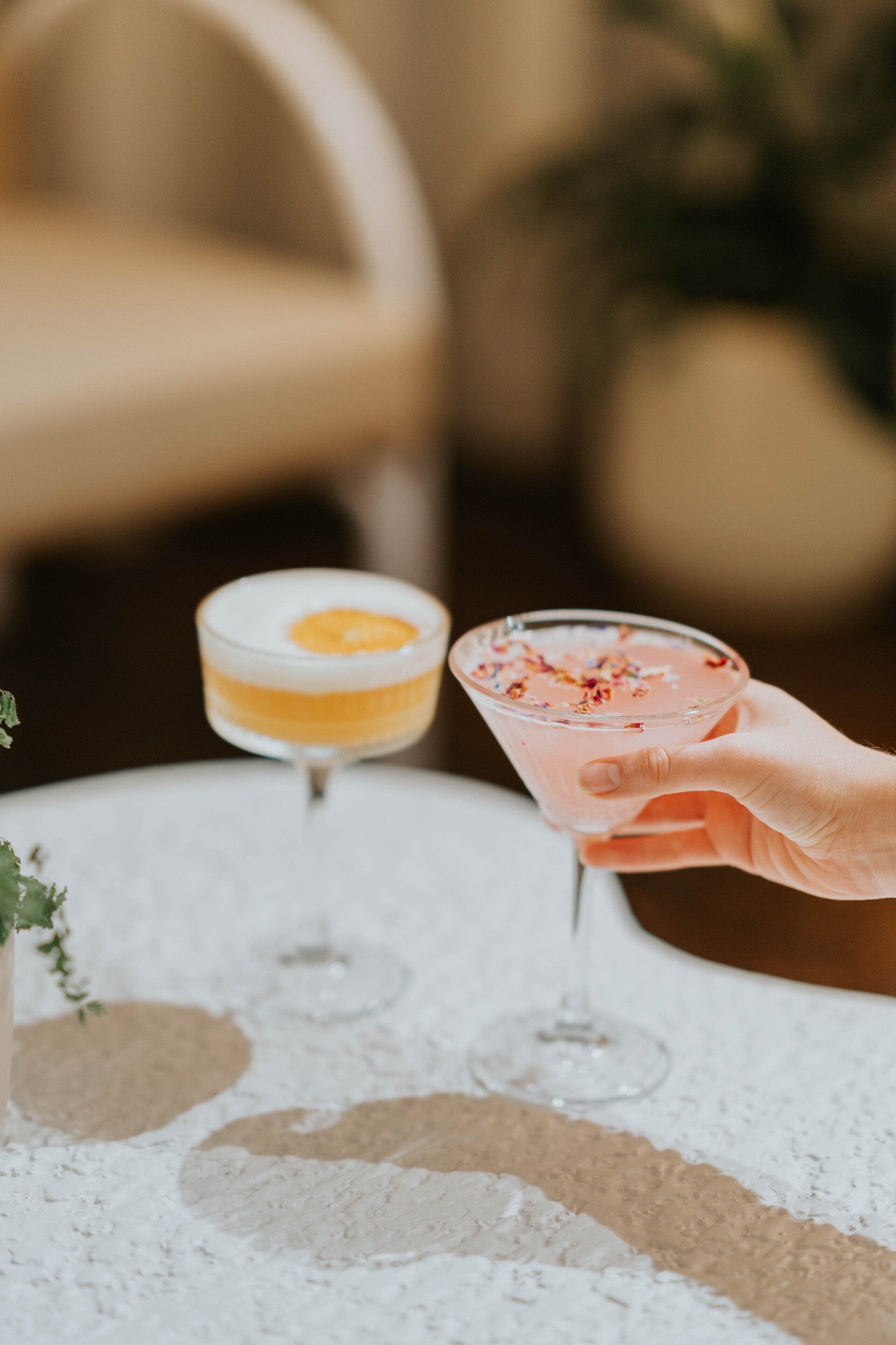 Hand holding a pink cocktail beside a white and yellow drink on a sunlit table
