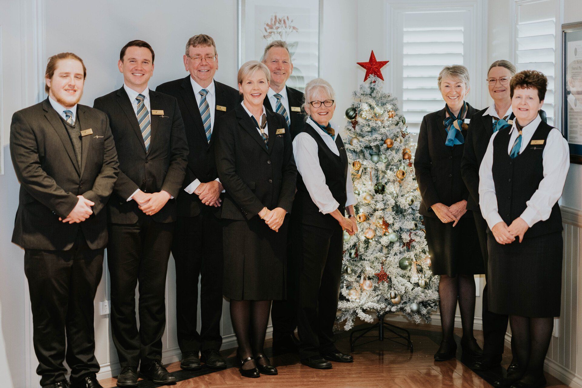 Nine people in formal attire standing beside a decorated Christmas tree indoors.