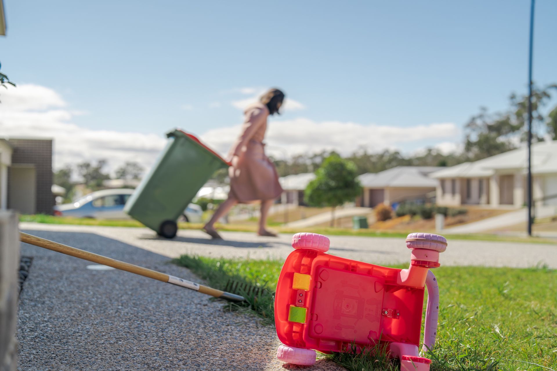 A tipped-over red toy car on a curb while a person pulls a green trash bin down a suburban street.