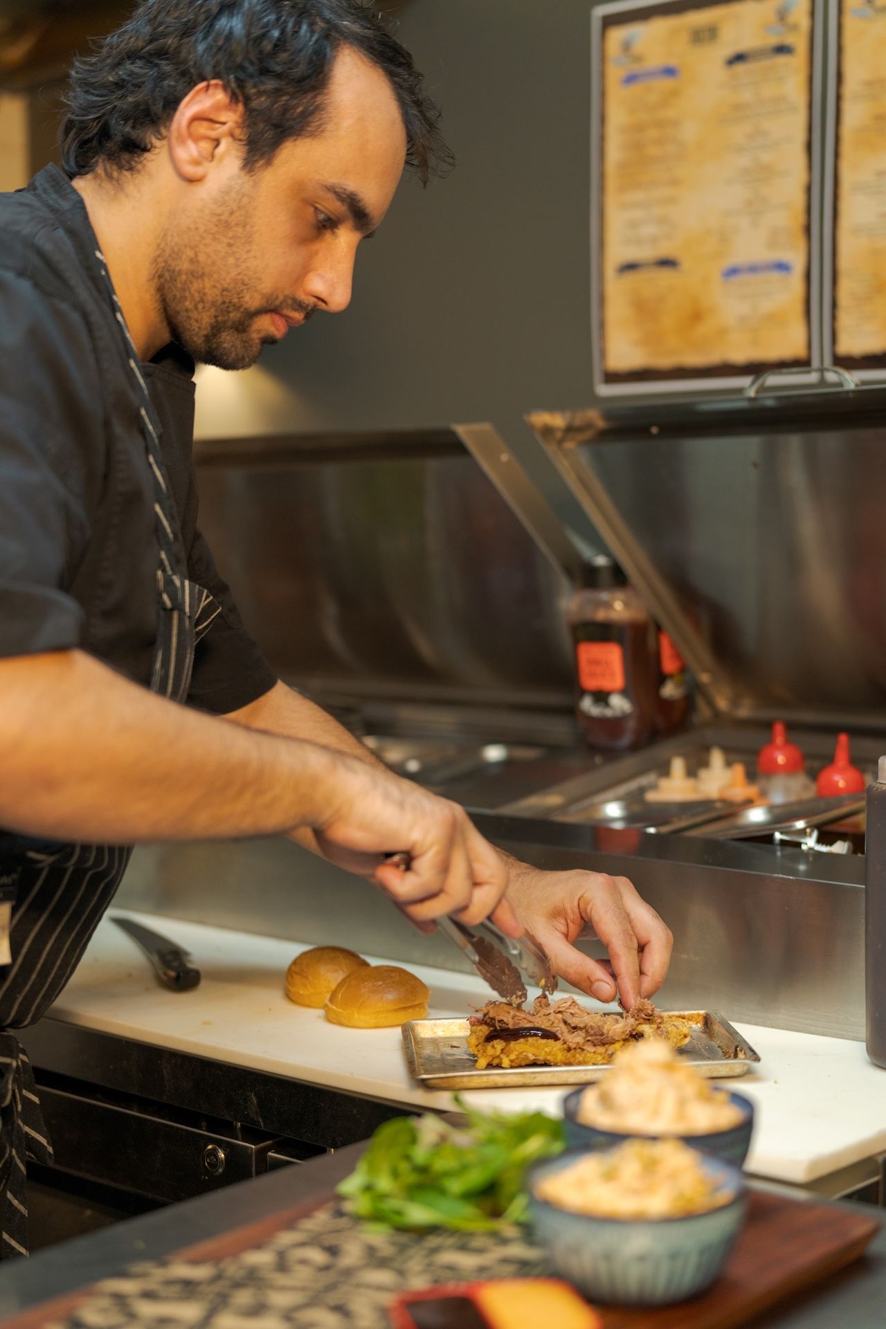 Chef plating food at a stainless-steel kitchen counter, with bowls of greens and ingredients nearby