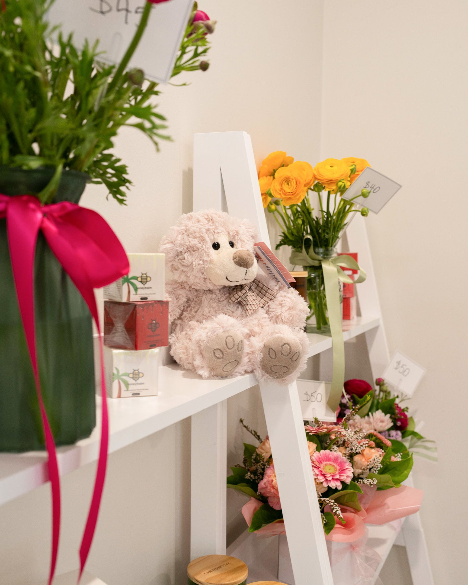 Teddy bear on a ladder shelf with flowers and gift baskets, decorated with pink ribbons.