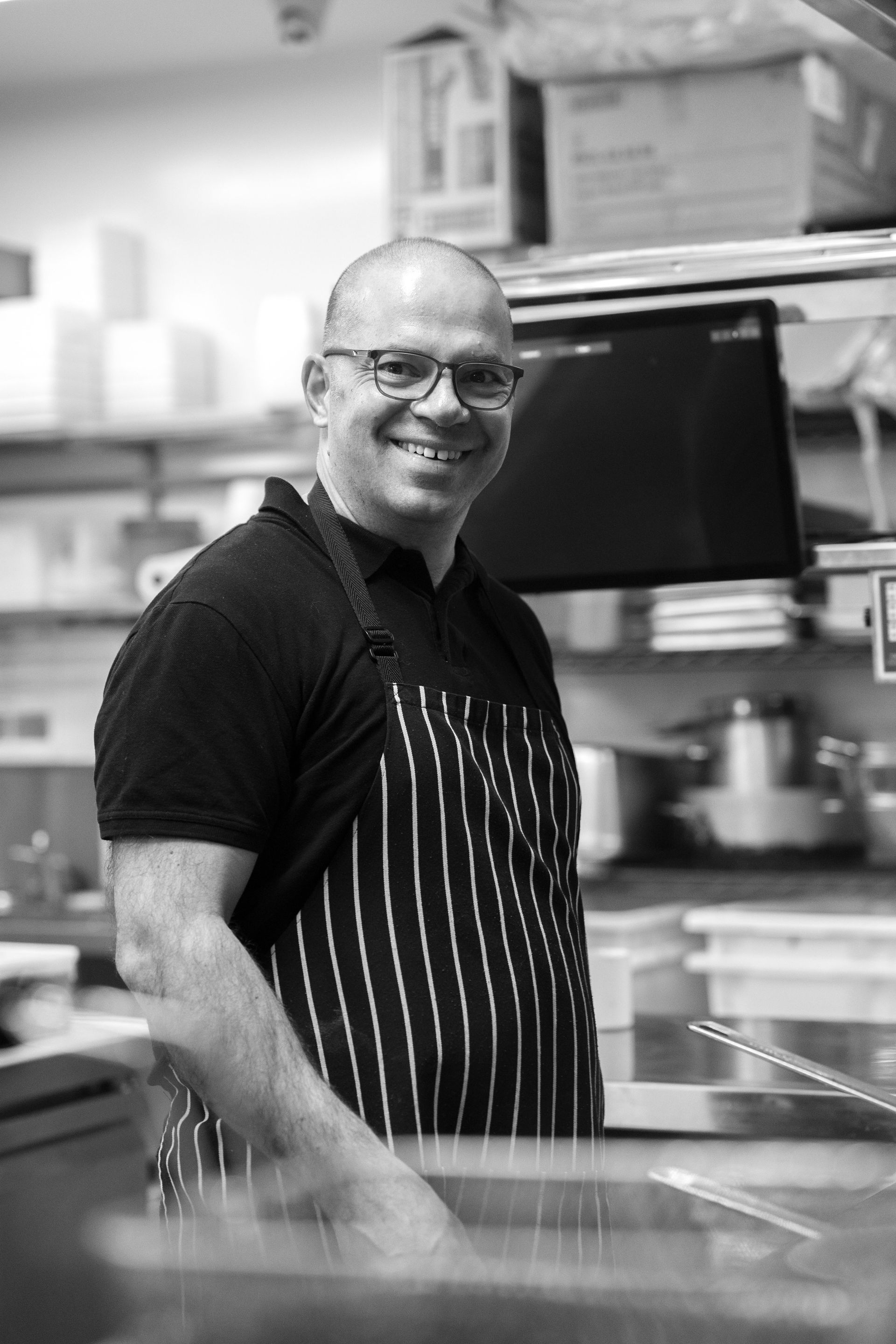 Smiling chef in striped apron standing in a kitchen, with shelves and a monitor behind him