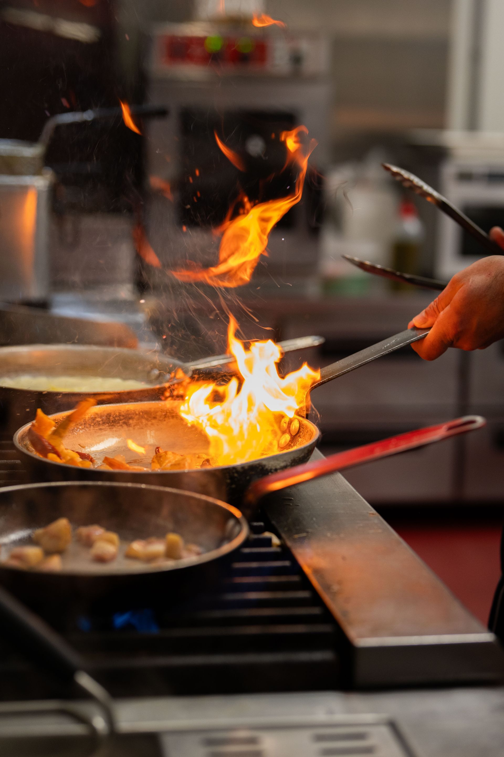 Chef tossing flaming food in a pan on a stovetop in a busy kitchen