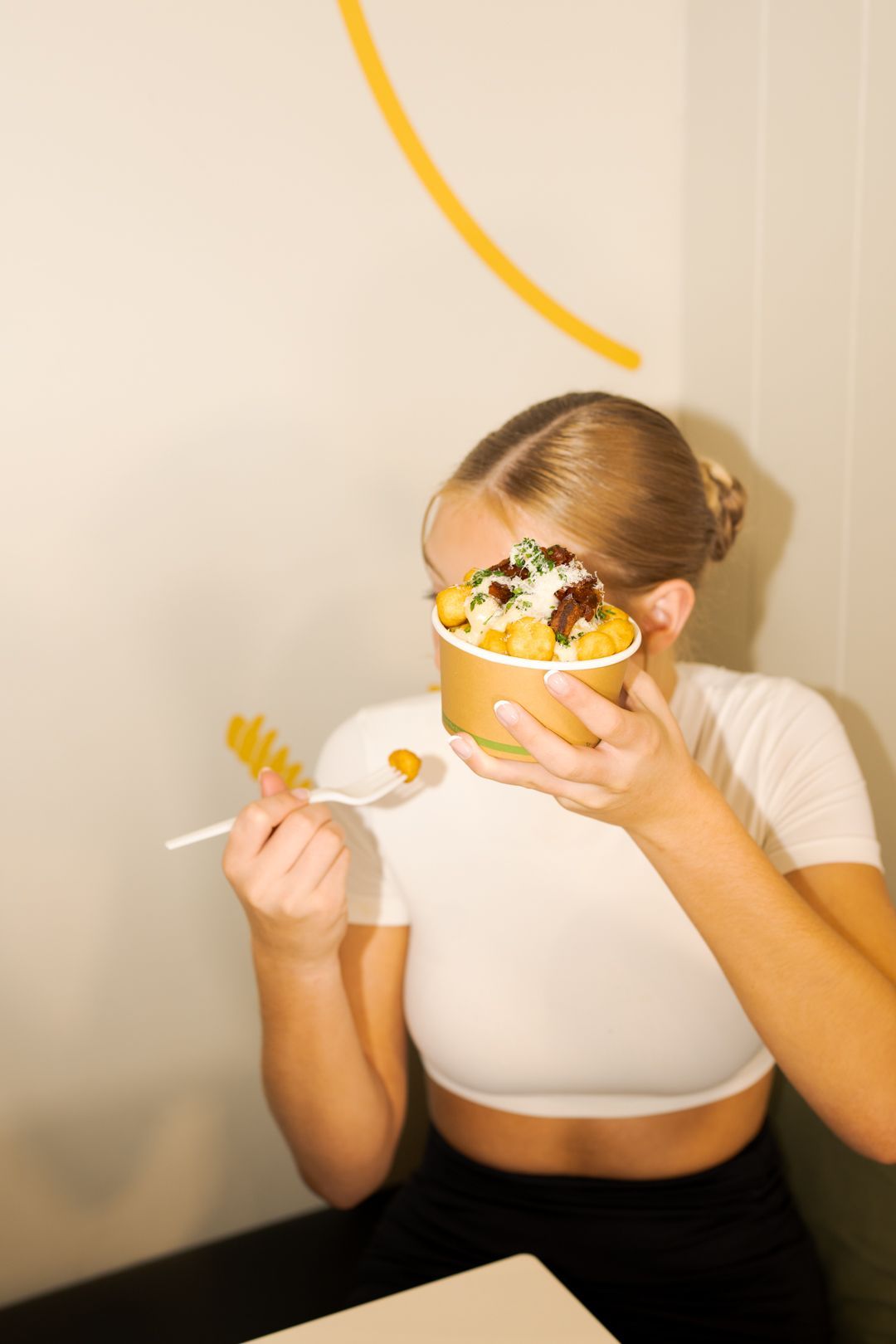 Person holding a dessert bowl and spoon, with whipped cream and fruit, against a light wall.