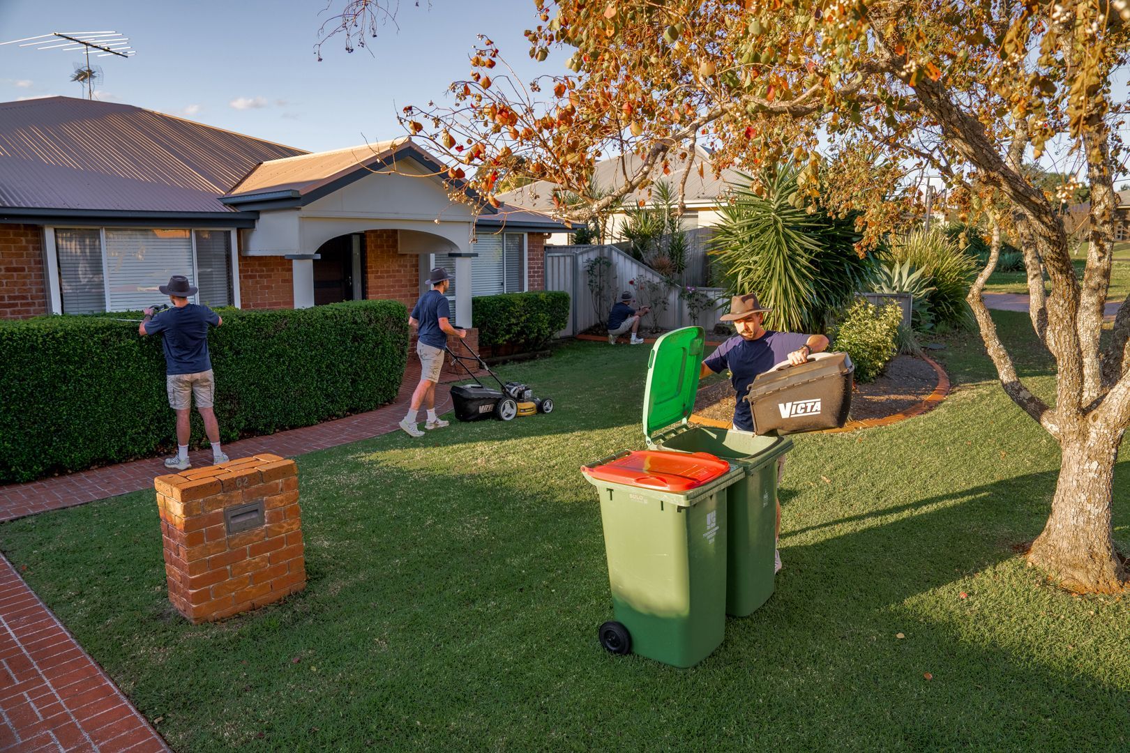 Workers loading green and black bins on a suburban lawn with a trash truck nearby