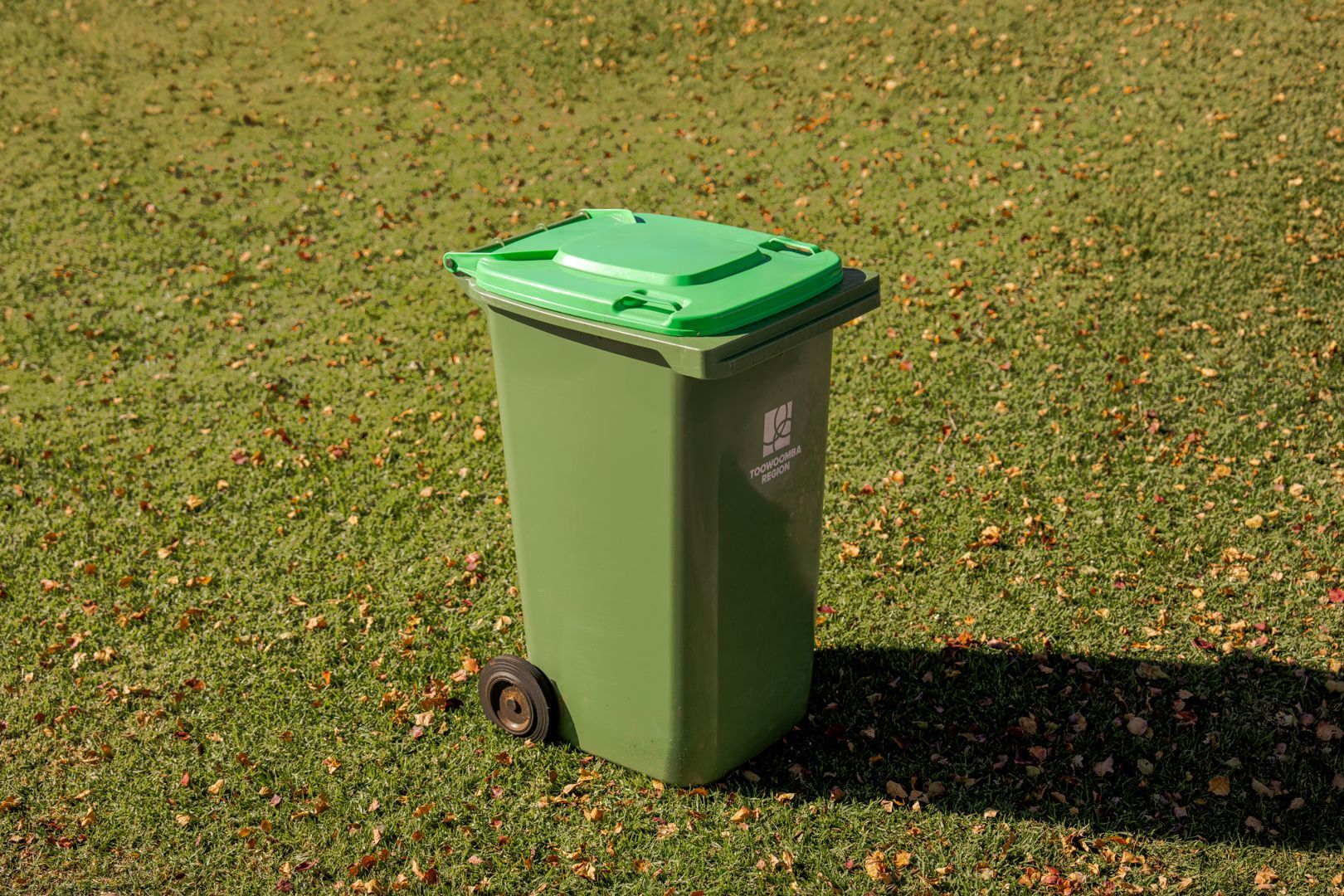 Green wheelie bin on a grassy lawn with scattered leaves.