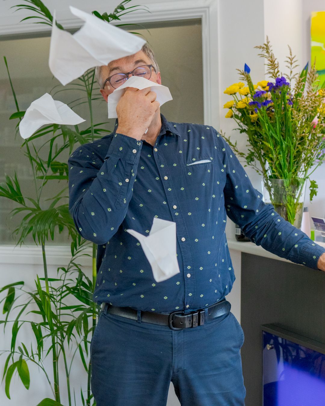 Man blowing tissues in a room, with several tissues flying around and flowers on a table nearby